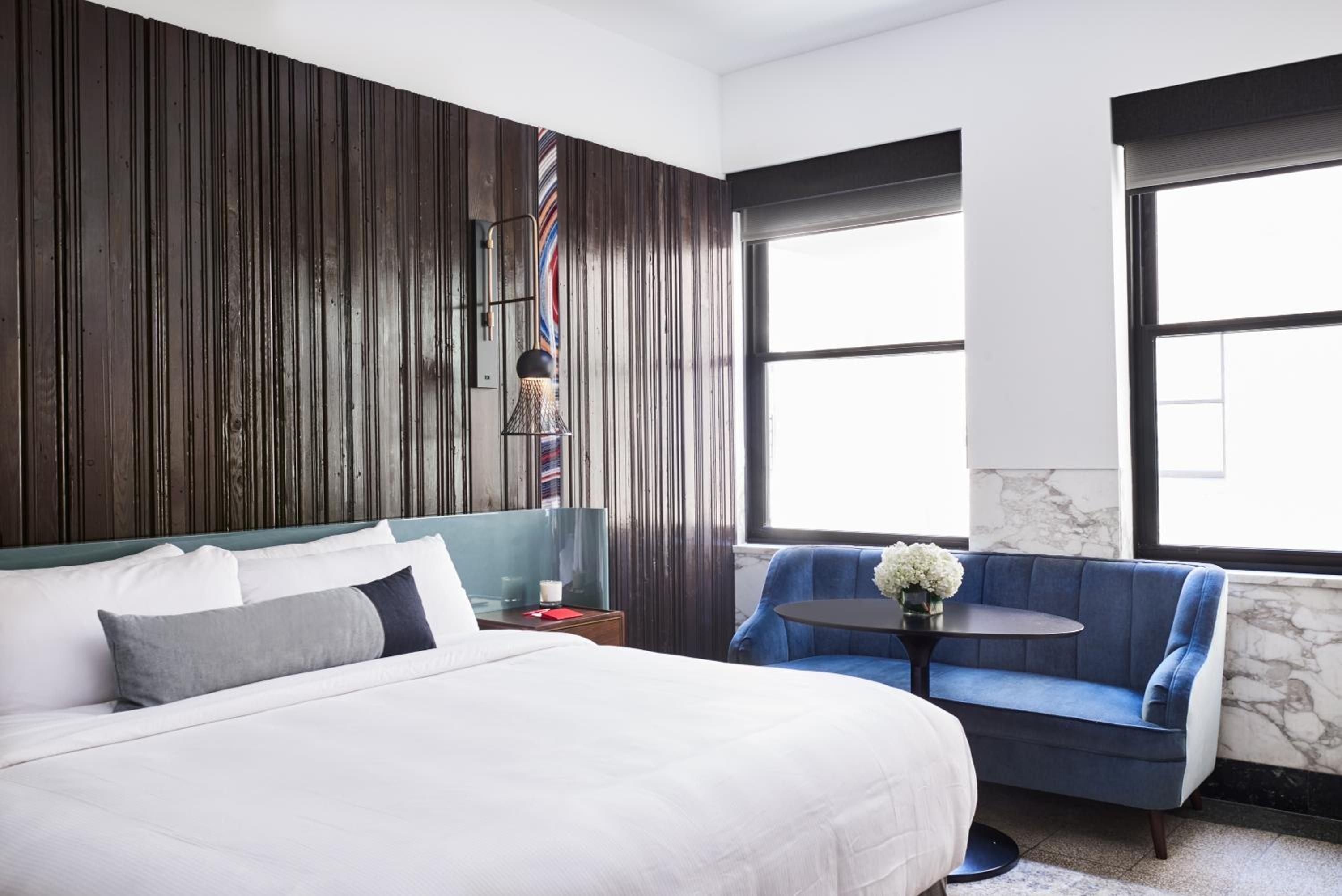 Vertical wood slats create dramatic shadows while plush blue velvet seating and crisp white linens balance industrial charm with luxurious comfort in this thoughtfully designed guest room.
