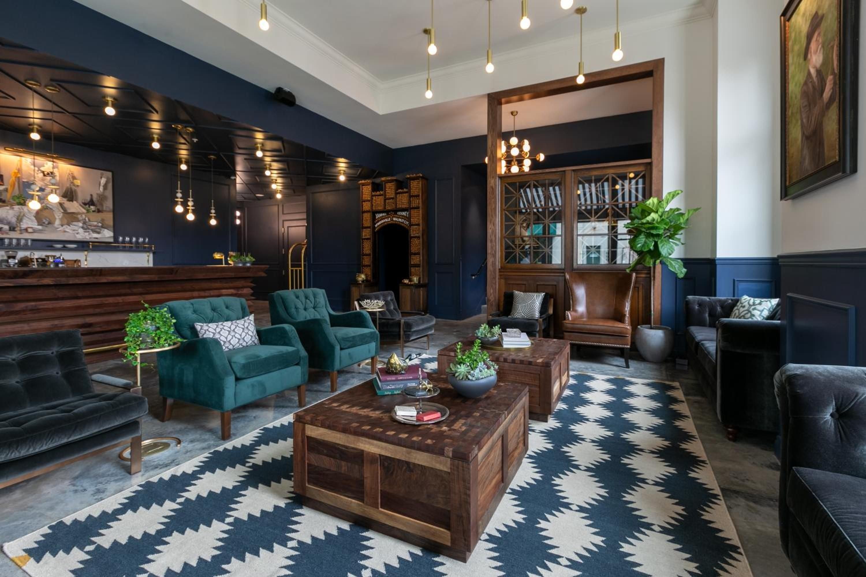 Rich navy walls and geometric carpets create an intimate gathering space where brass pendant lights cast warm glows over plush teal armchairs and handcrafted walnut coffee tables.
