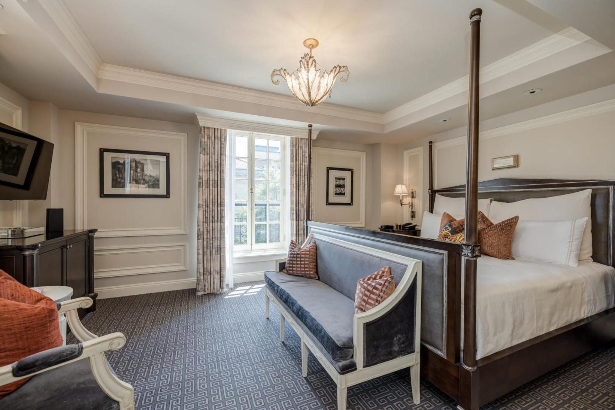 Elegant four-poster bed anchors this sophisticated guest room, where warm neutrals, geometric carpeting, and crystal chandelier create an atmosphere of refined Southern hospitality.