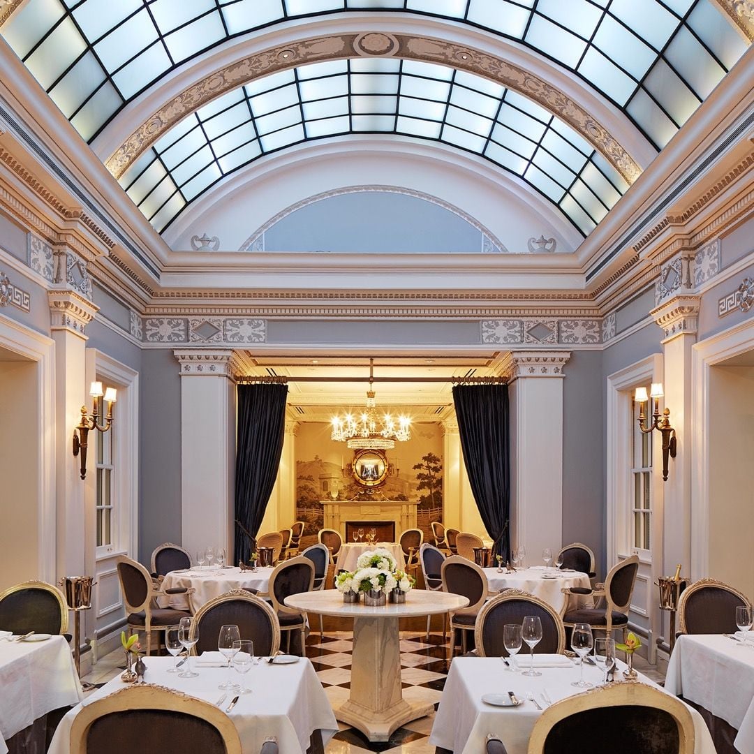 Beneath soaring glass ceilings and ornate moldings, intimate tables await in this neoclassical dining room where crystal chandeliers cast golden light across black and white checkered floors.