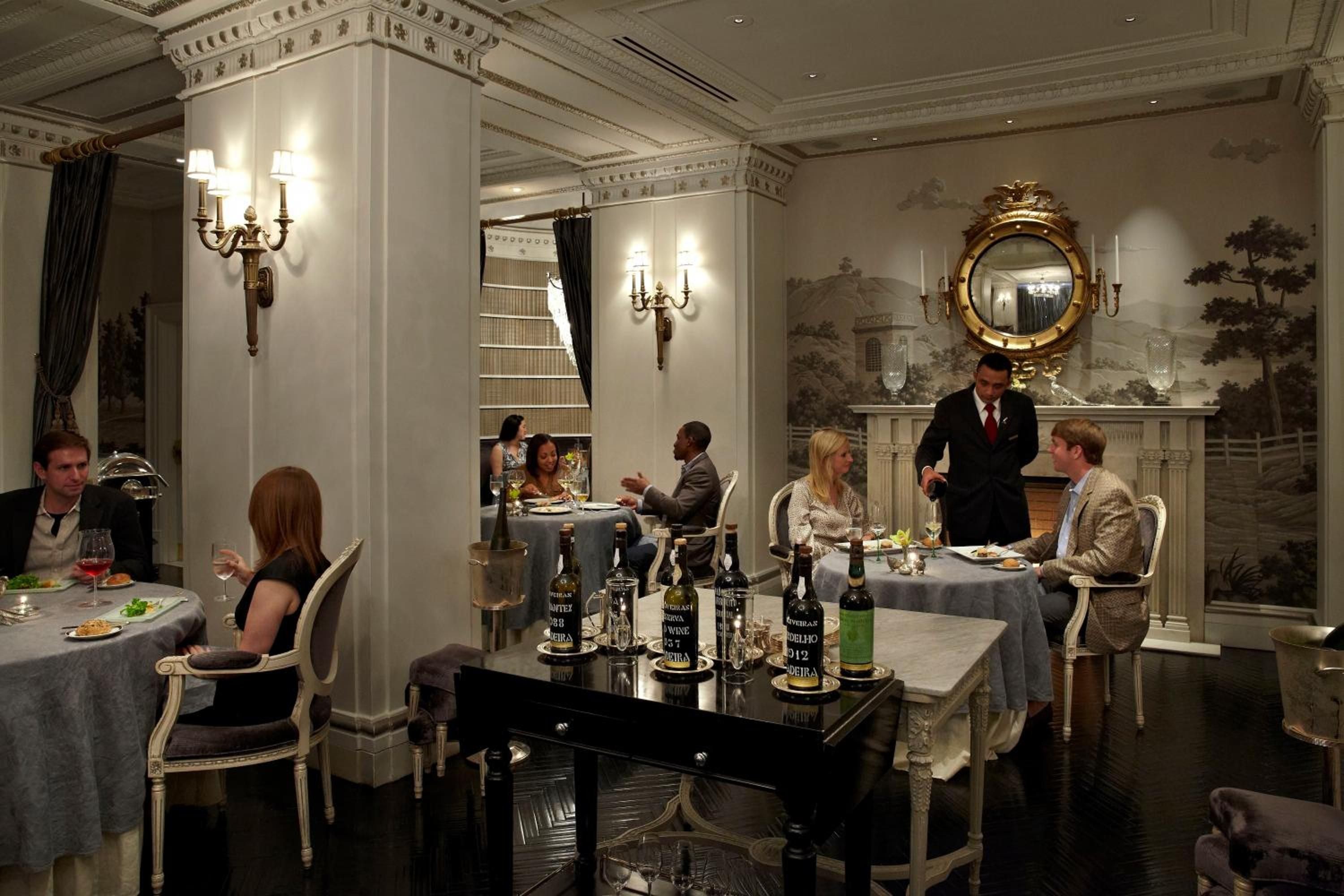 Crystal sconces cast warm light across intimate dining tables while a tuxedoed server tends to guests in this elegant restaurant's classic European-inspired setting.
