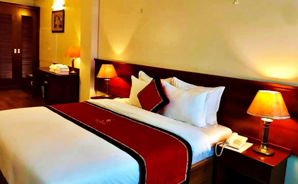 Warm amber lighting bathes the elegant guest room, where rich mahogany furnishings and crisp white linens create an inviting sanctuary in Vietnam's bustling capital.