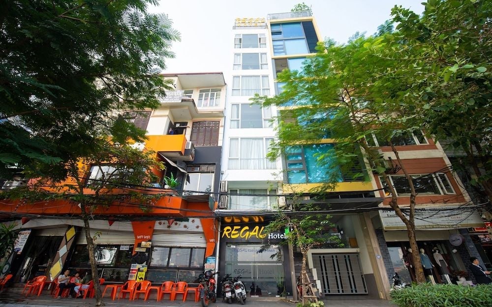 Gleaming white facade rises above bustling Hanoi streets, where orange café chairs spill onto sidewalks and tropical greenery frames this sleek urban retreat.