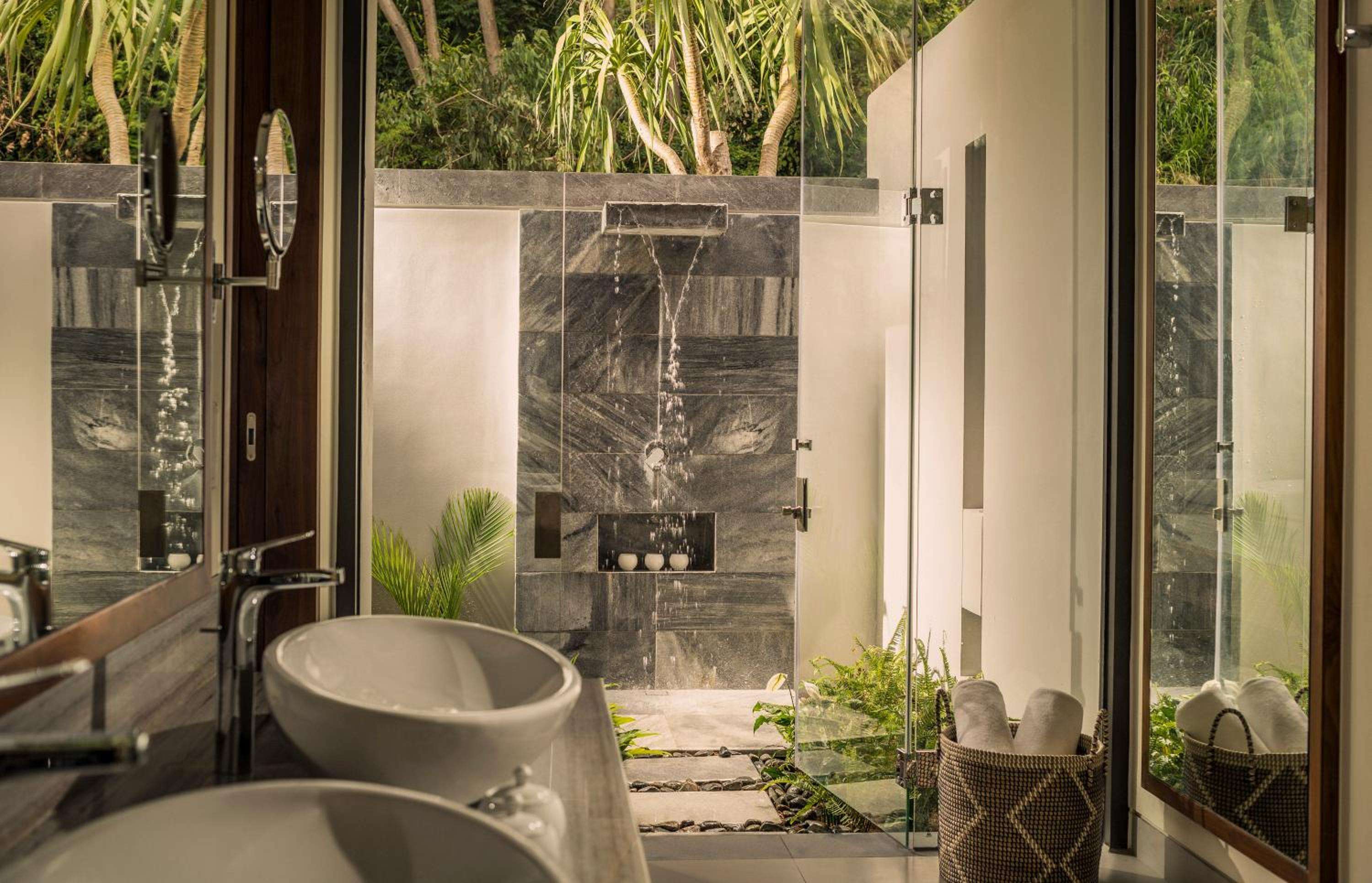 Natural stone walls frame an open-air rain shower while floor-to-ceiling glass dissolves boundaries between marble vanity and lush tropical garden beyond.