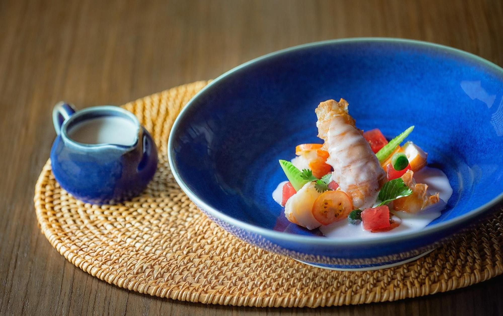 Artfully plated Vietnamese cuisine arrives in handcrafted blue ceramics, where delicate prawns and vibrant vegetables create an intimate culinary moment against woven textures.