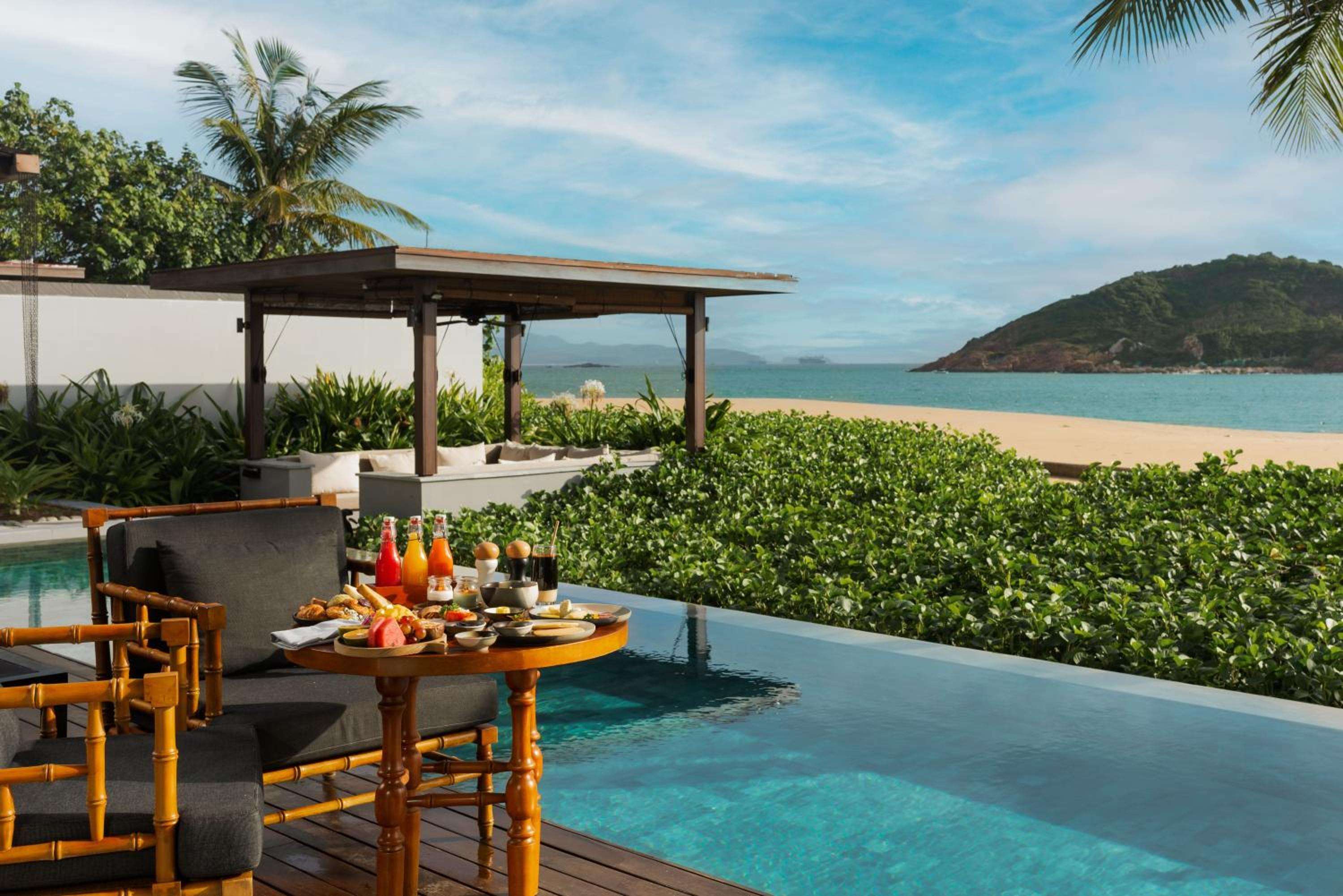 Morning light dances across an infinity pool where breakfast awaits, bamboo chairs positioned perfectly to frame Quy Nhon's pristine coastline and verdant hills beyond.
