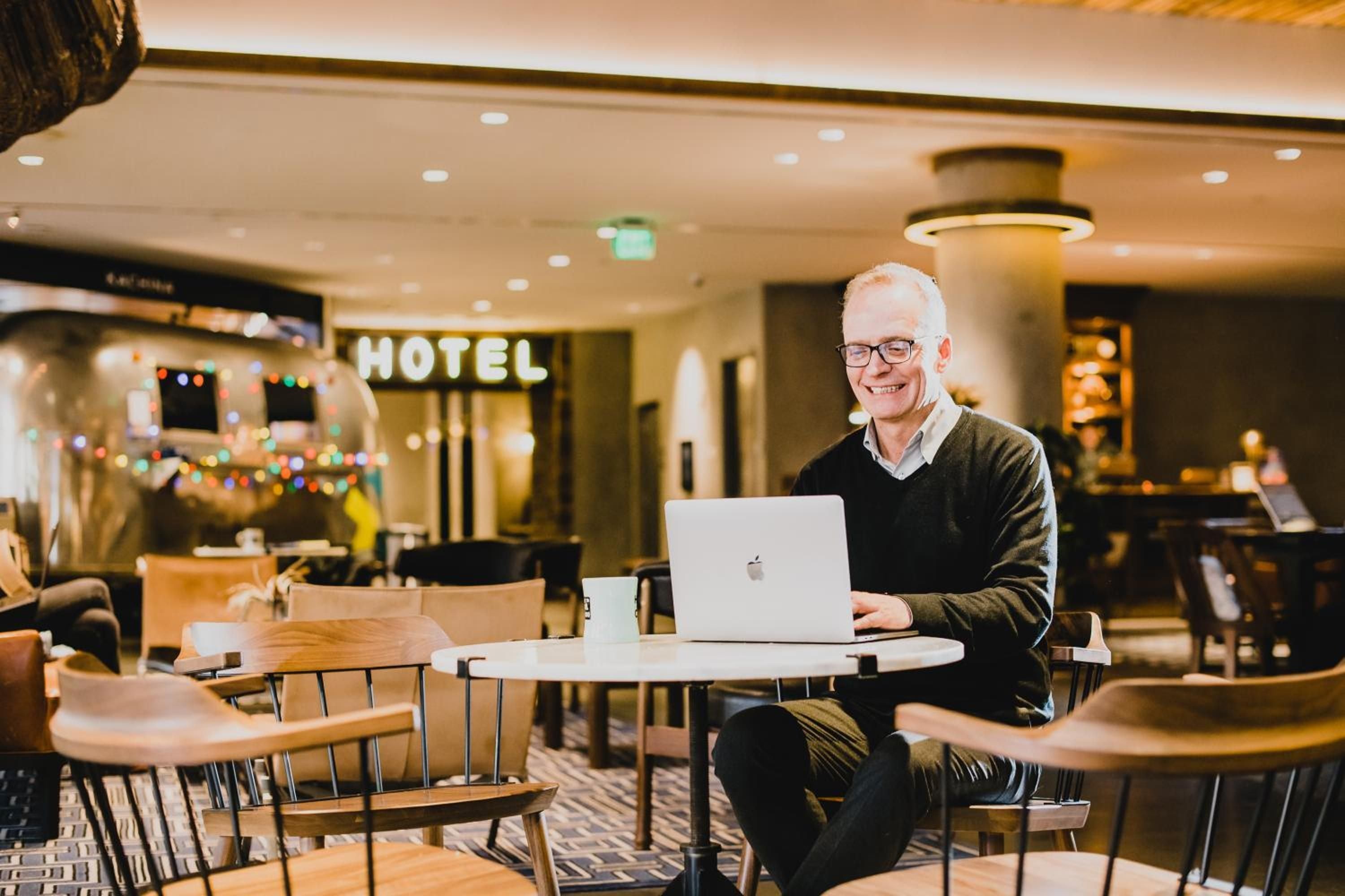 Silver-haired gentleman finds his workflow in the hotel's airy common space, where industrial chic meets comfort beneath warm pendant lighting and exposed beams.