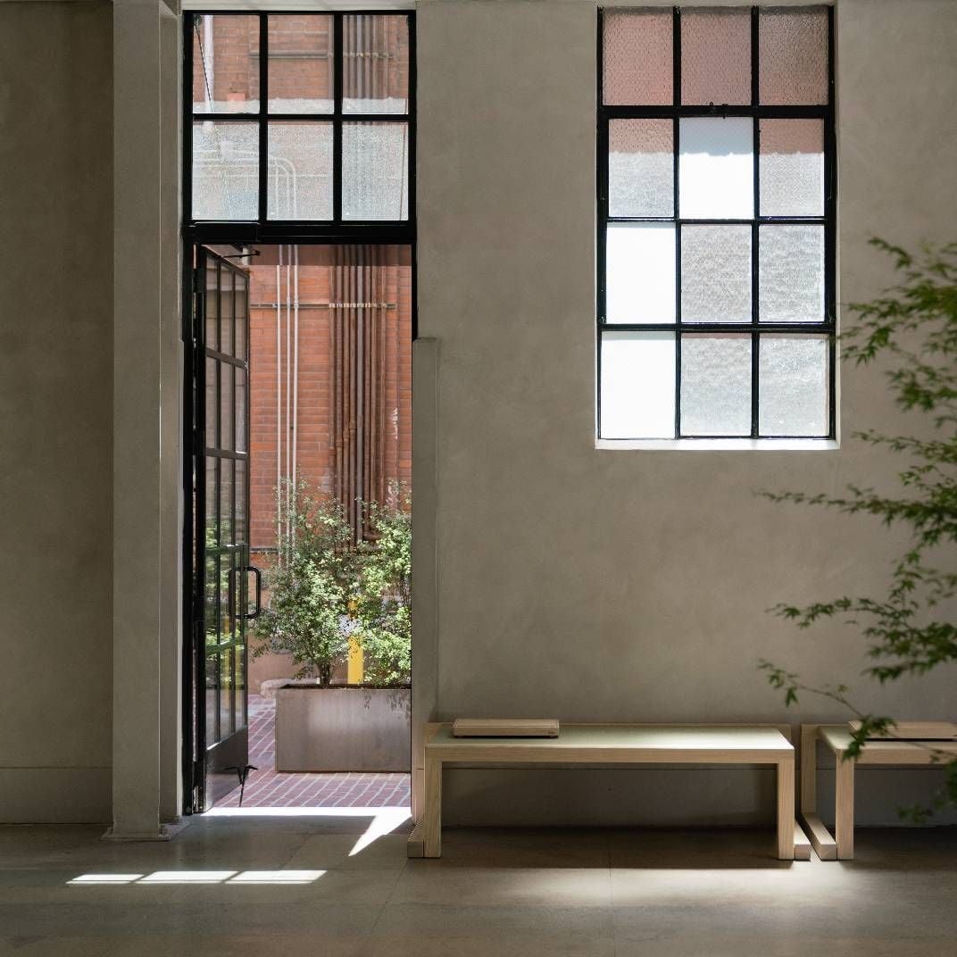 Sunlight streams through industrial-framed windows onto minimalist wooden benches, while glimpses of brick courtyards hint at hidden urban sanctuaries beyond.