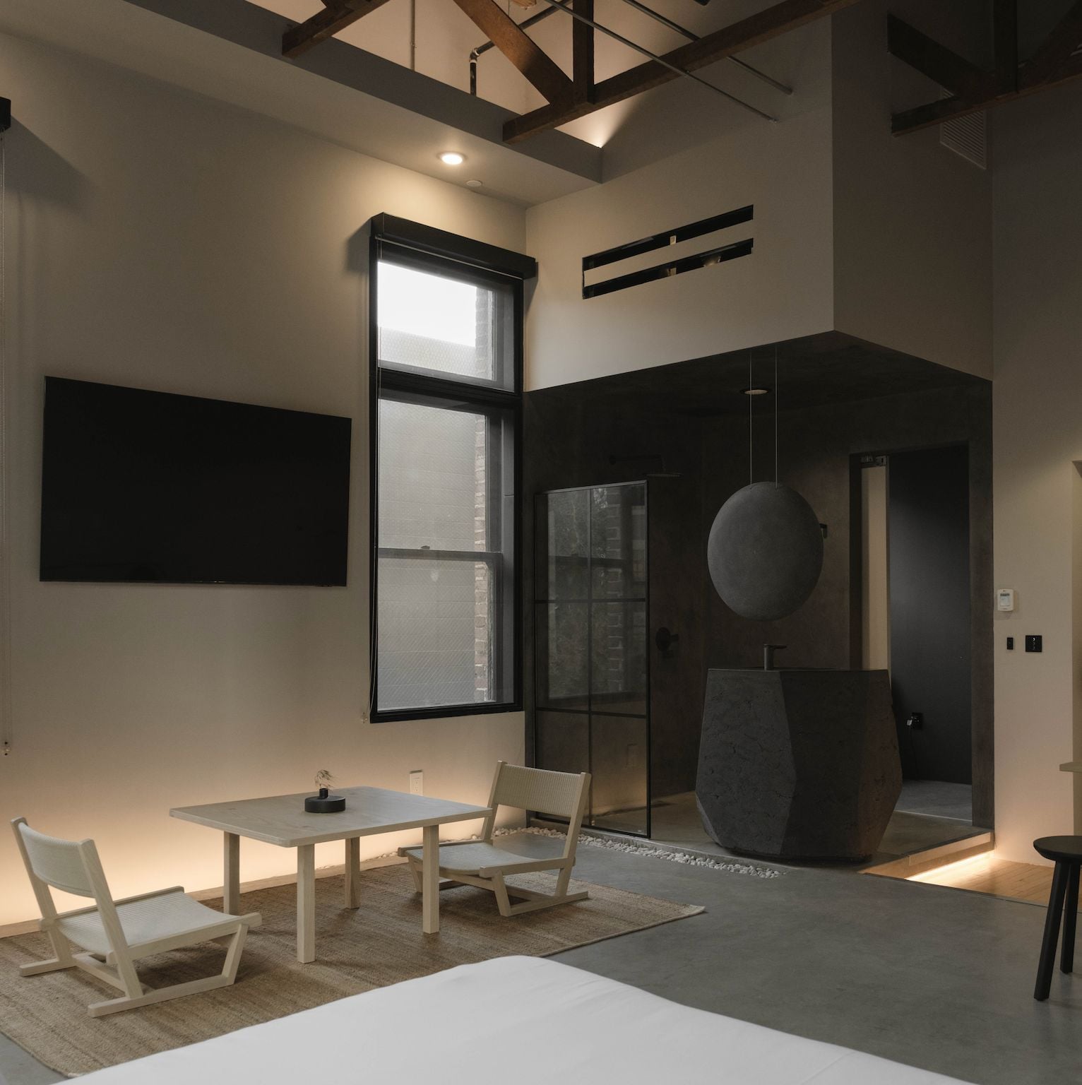 Exposed wooden beams frame a serene sanctuary where minimalist Japanese design meets industrial chic, featuring a sculptural soaking tub and floor-to-ceiling glass shower enclosure.