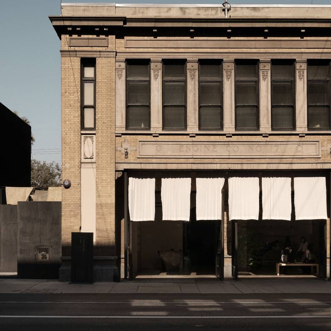 Warm afternoon light bathes the historic brick facade, where white curtains flutter in open garage-style doors, creating an intriguing blend of industrial heritage and contemporary hospitality.