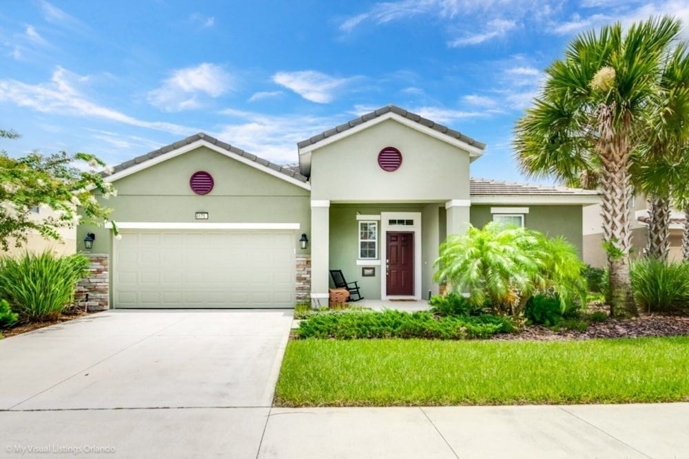 Sage-green stucco walls and burgundy accents create curb appeal at this modern Florida villa, where tropical landscaping frames a welcoming covered entry.