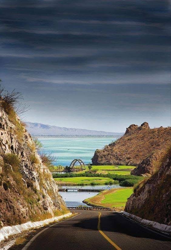Desert highway cuts through volcanic terrain toward emerald waters of Danzante Bay, where rugged cliffs frame golf greens and the Sea of Cortez stretches endlessly.