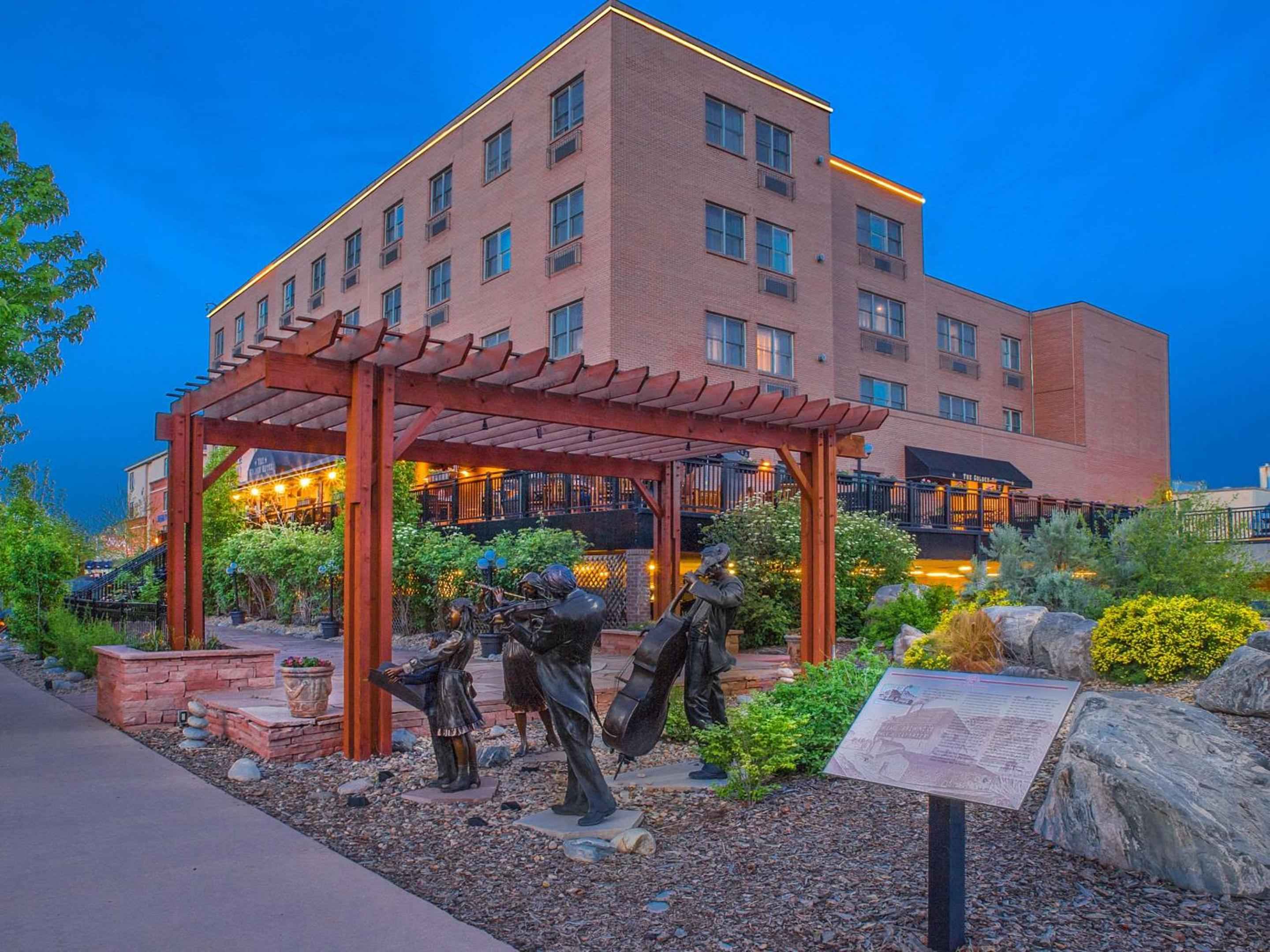 Bronze sculptures of weary travelers rest beneath a timber pergola, while the hotel's warm brick facade glows against Colorado's impossibly blue twilight sky.