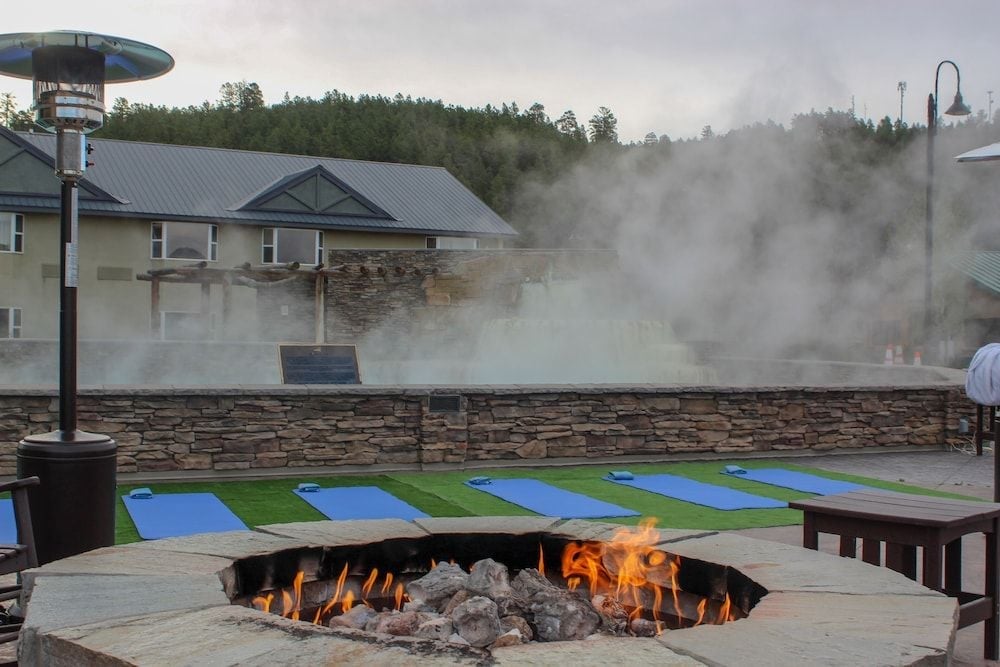 Steam rises from natural hot springs while a crackling fire pit anchors the yoga deck, creating an atmospheric evening retreat against the forested mountainside.