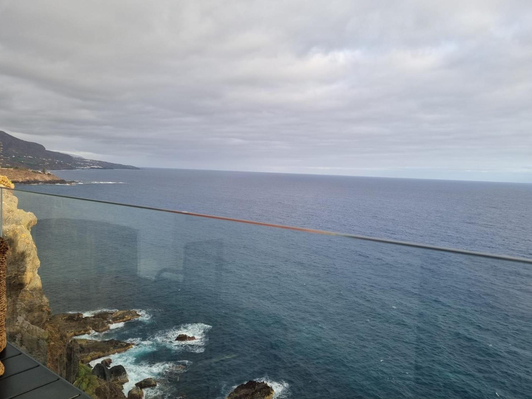 From the glass-railed terrace, dramatic coastline unfolds beneath moody Atlantic skies, where volcanic cliffs meet cerulean waters in an endless dance of elements.