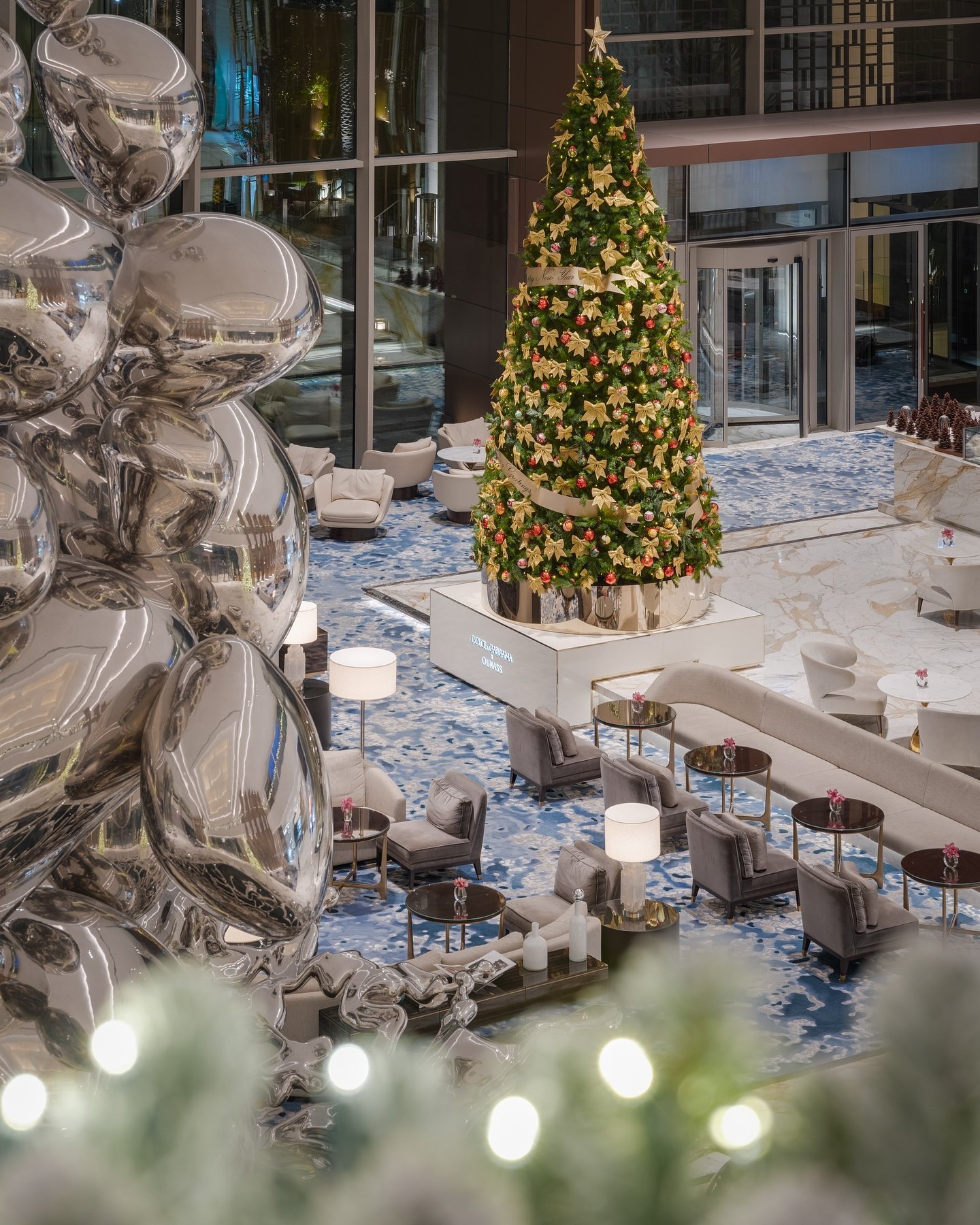 Metallic balloon sculptures catch light beside a towering Christmas tree in this sophisticated lobby, where marble floors and plush seating create an unexpected festive elegance.