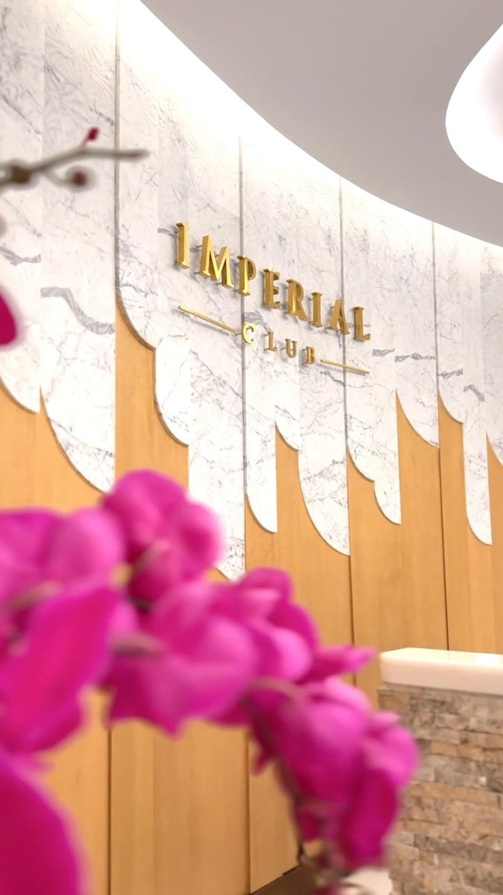 Golden lettering gleams against marble-veined walls where warm wood meets white stone, while vibrant orchids add unexpected pops of fuchsia to the sophisticated entrance.