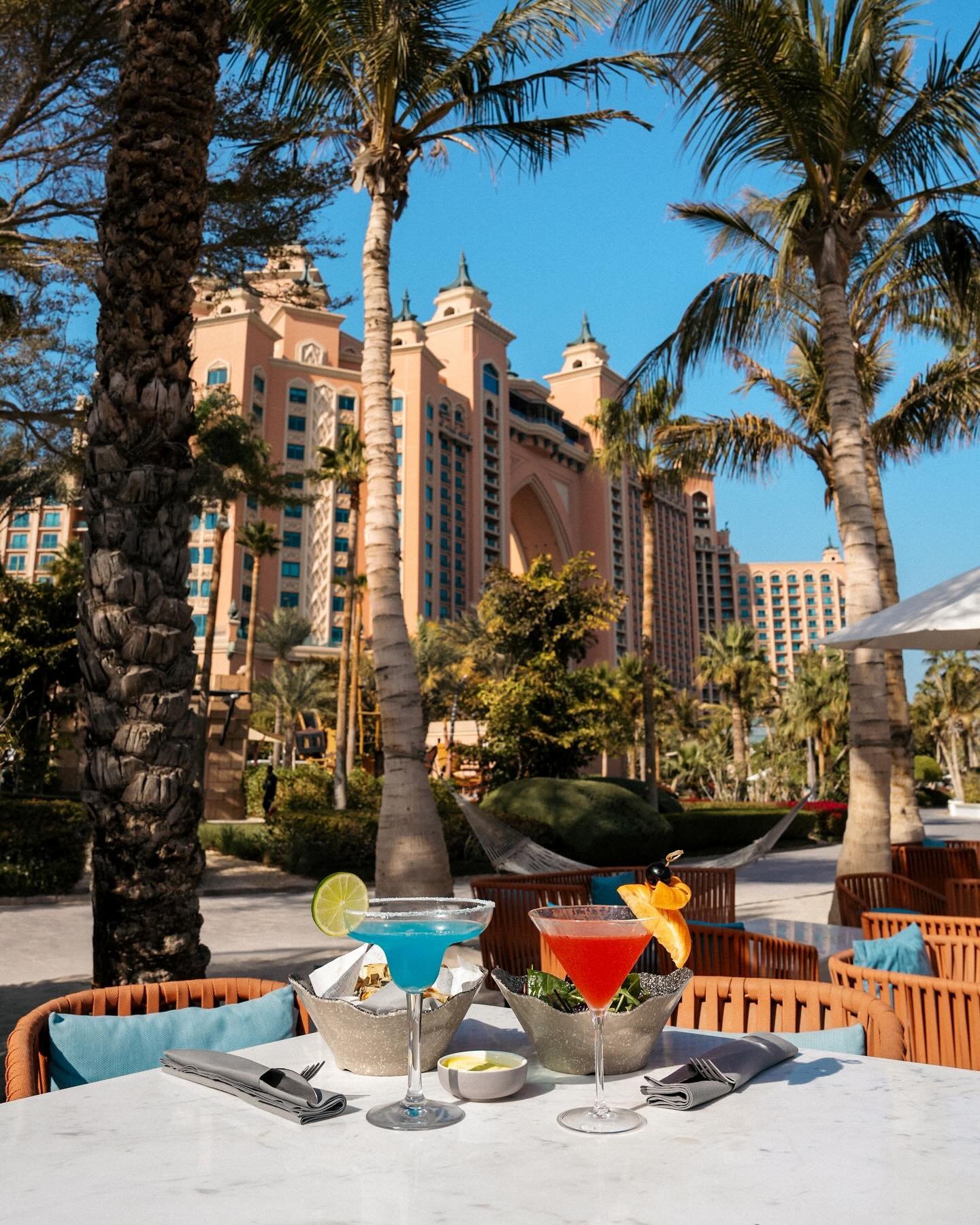 Tropical cocktails catch golden hour light at this poolside table, where Atlantis Resort's iconic pink towers create a dreamy backdrop through swaying palms.