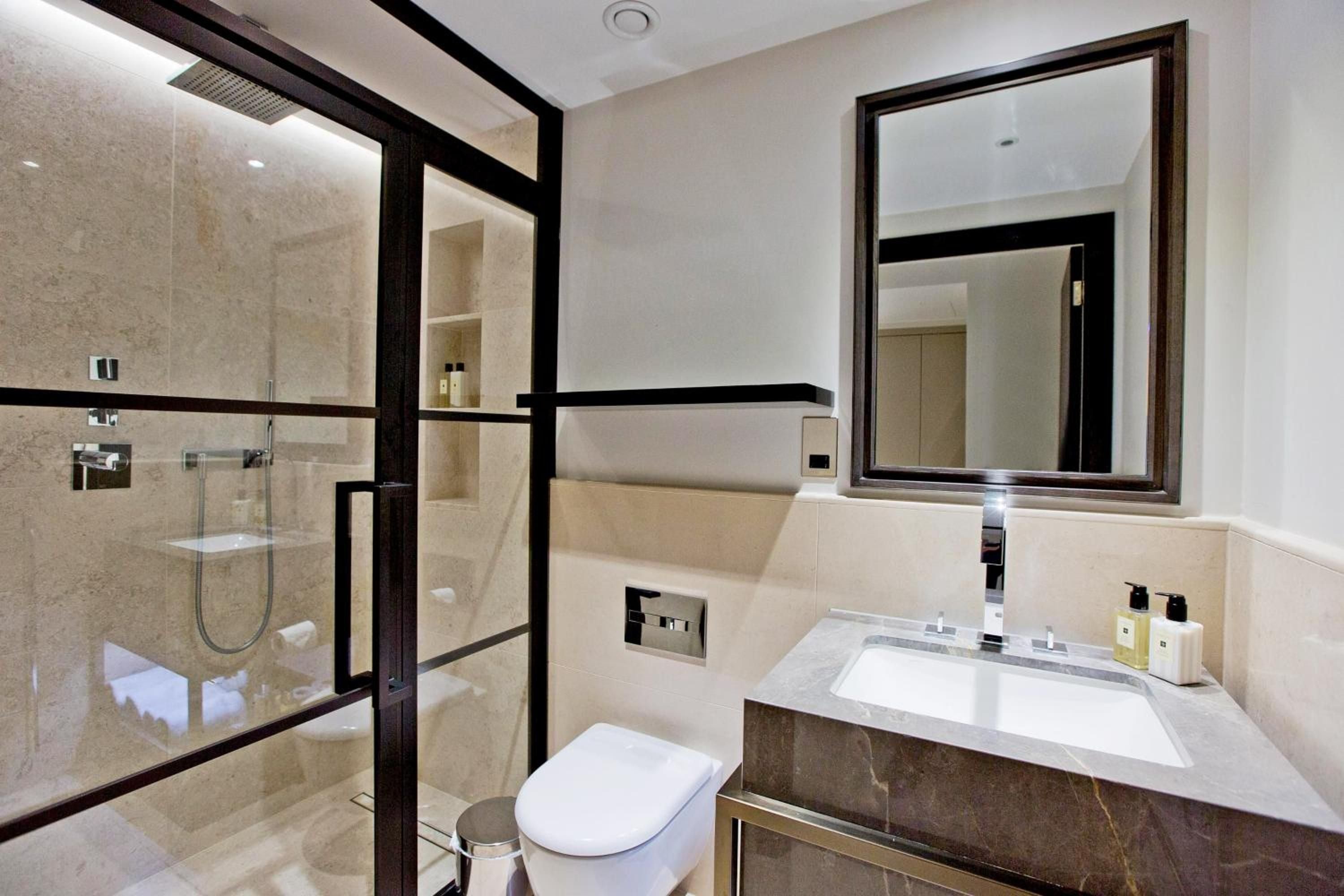 Sleek black-framed glass dividers separate the marble-clad shower from sculptural basin, where warm lighting highlights the sophisticated interplay of stone and chrome fixtures.