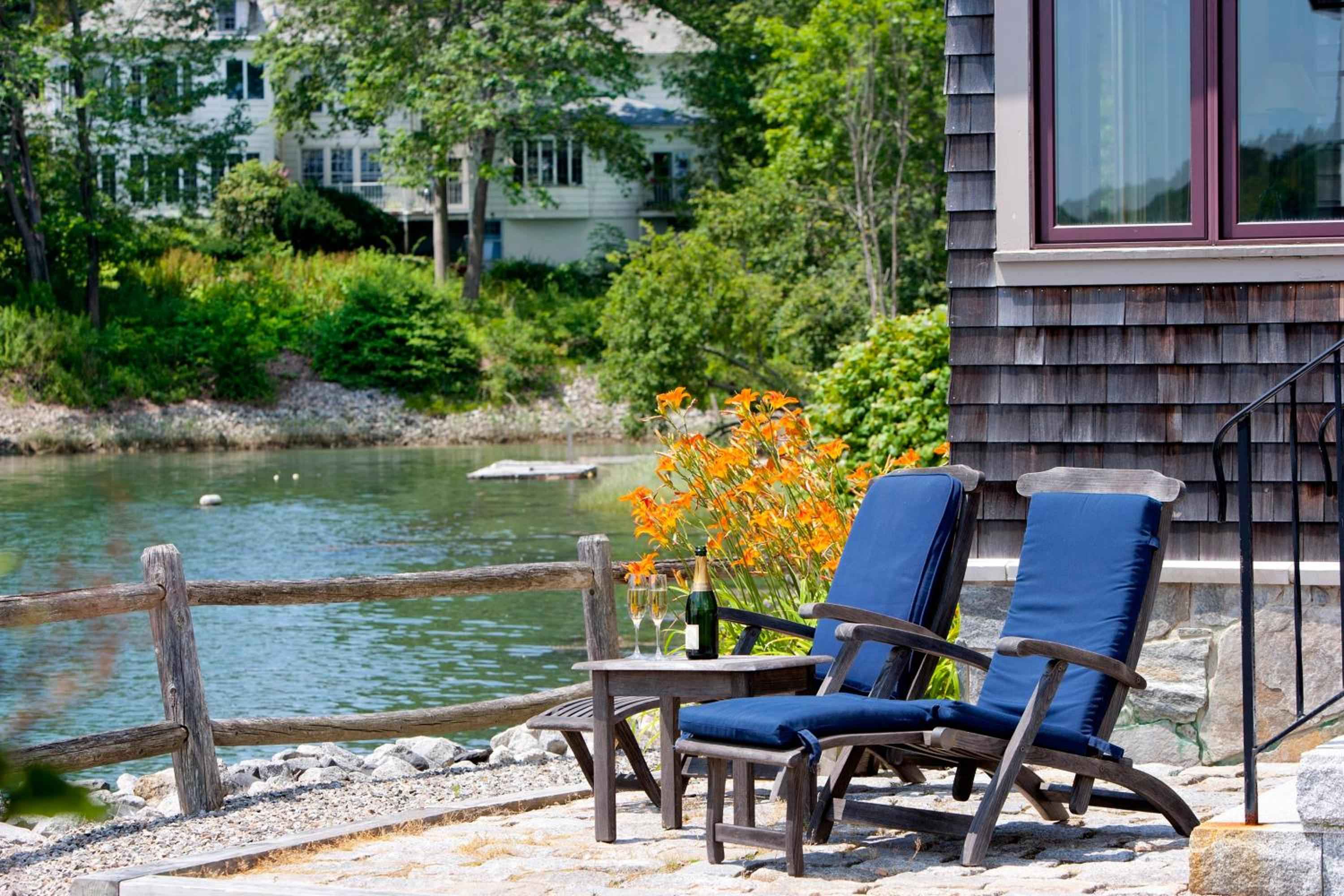 Blue-cushioned loungers overlook tranquil harbor waters, where weathered cedar shingles and orange day lilies create an quintessentially Maine coastal retreat.