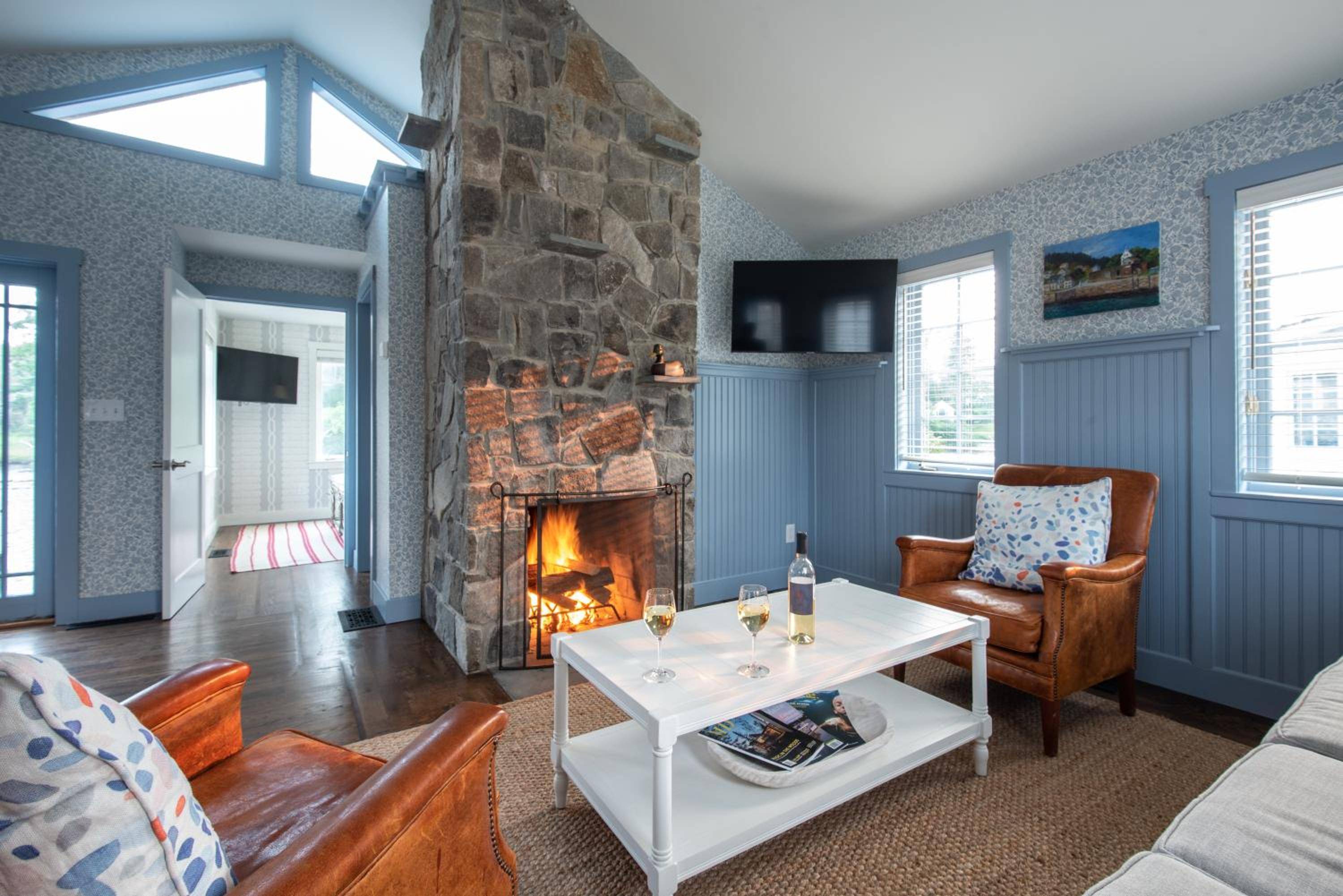 Stone fireplace crackles warmly in this coastal retreat's living area, where blue-washed walls and leather armchairs create an intimate New England sanctuary.