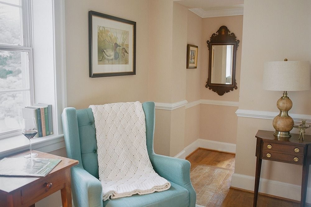 Soft afternoon light filters through tall windows onto a turquoise wingback chair, where cable-knit throws and vintage botanical prints create an inviting reading nook.