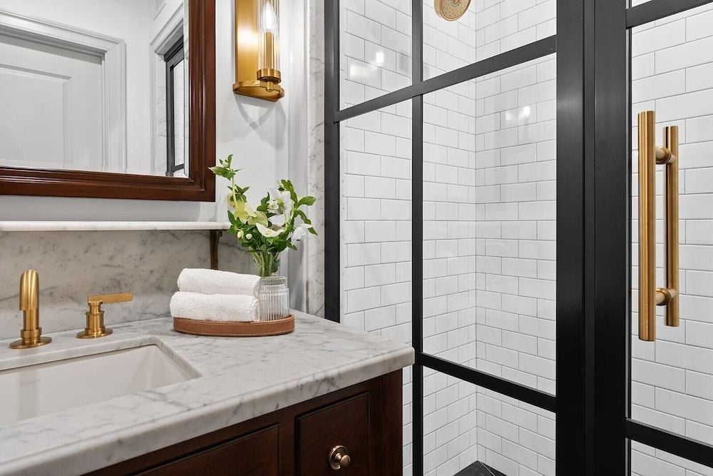 Brass fixtures gleam against Carrara marble while black-framed glass doors reveal subway-tiled showers, creating an elegant study in classic contrasts.