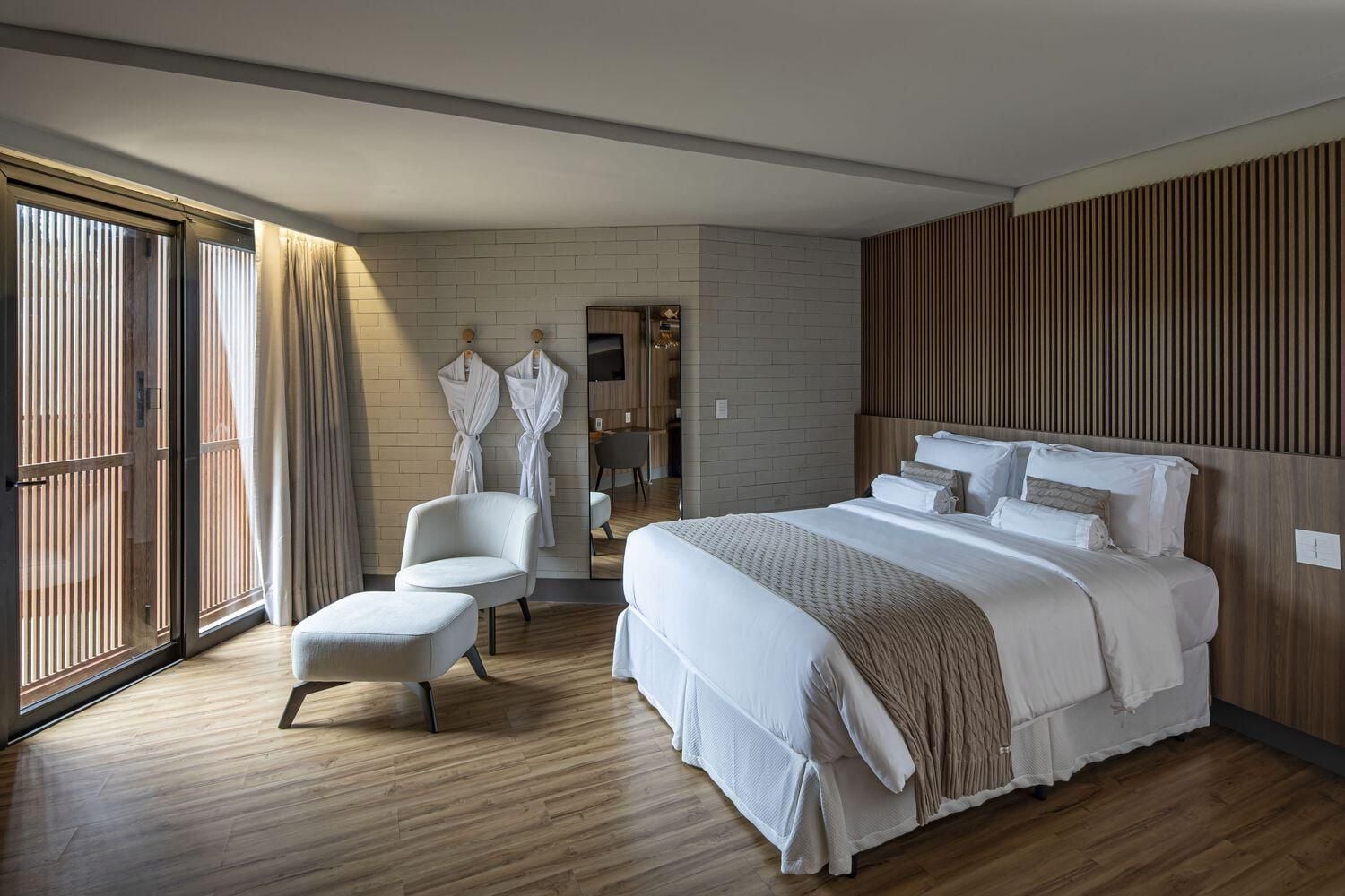 Warm sunlight filters through vertical slats, casting golden patterns across polished hardwood floors where crisp white linens meet rich wood paneling in perfect harmony.