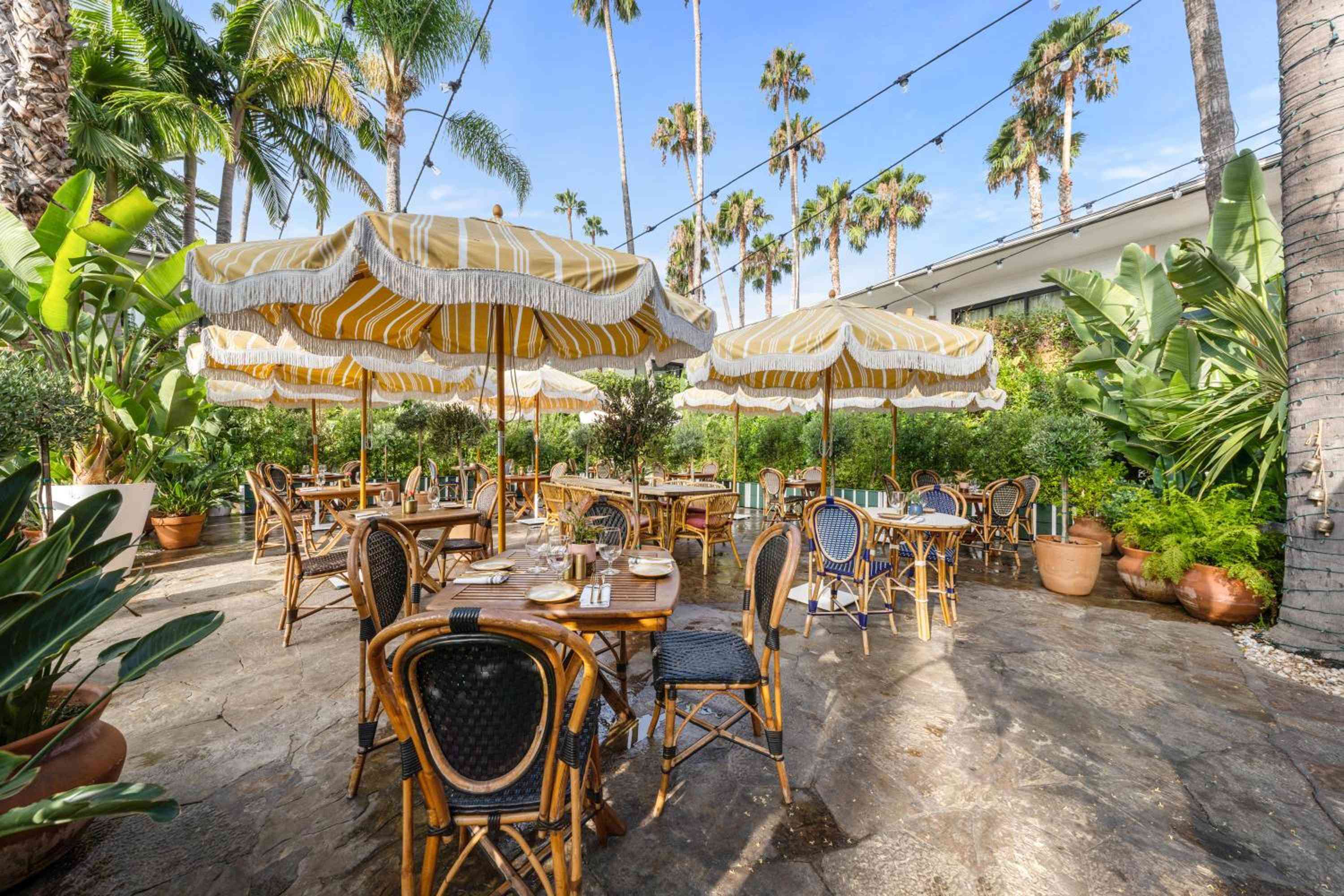 Striped umbrellas cast golden shadows over rattan bistro chairs, where palm fronds whisper overhead and terracotta planters frame this enchanting courtyard dining escape.
