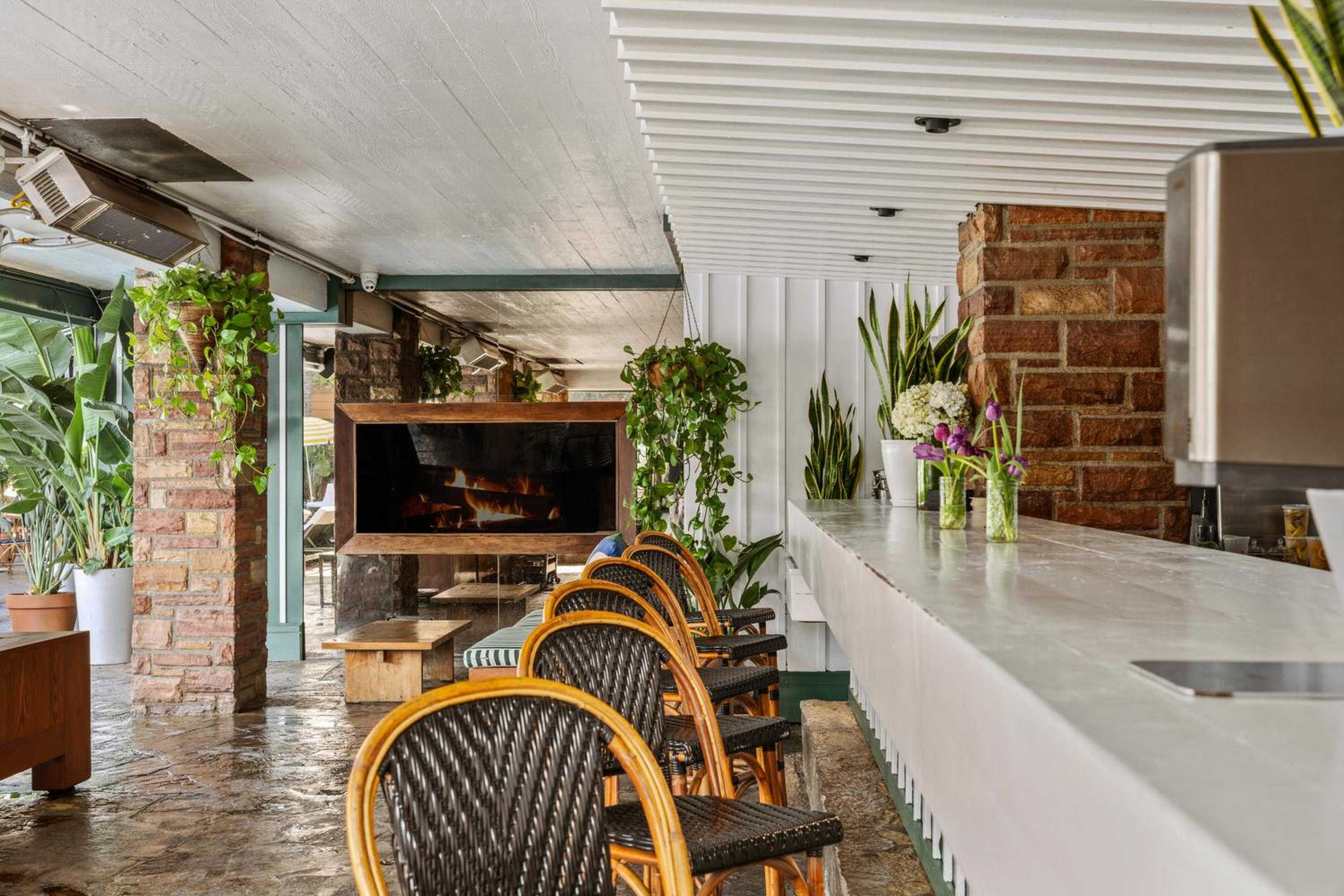 Wicker bistro chairs line the marble bar where tropical plants cascade from exposed brick walls, creating an intimate greenhouse-like sanctuary with flickering fireplace warmth.