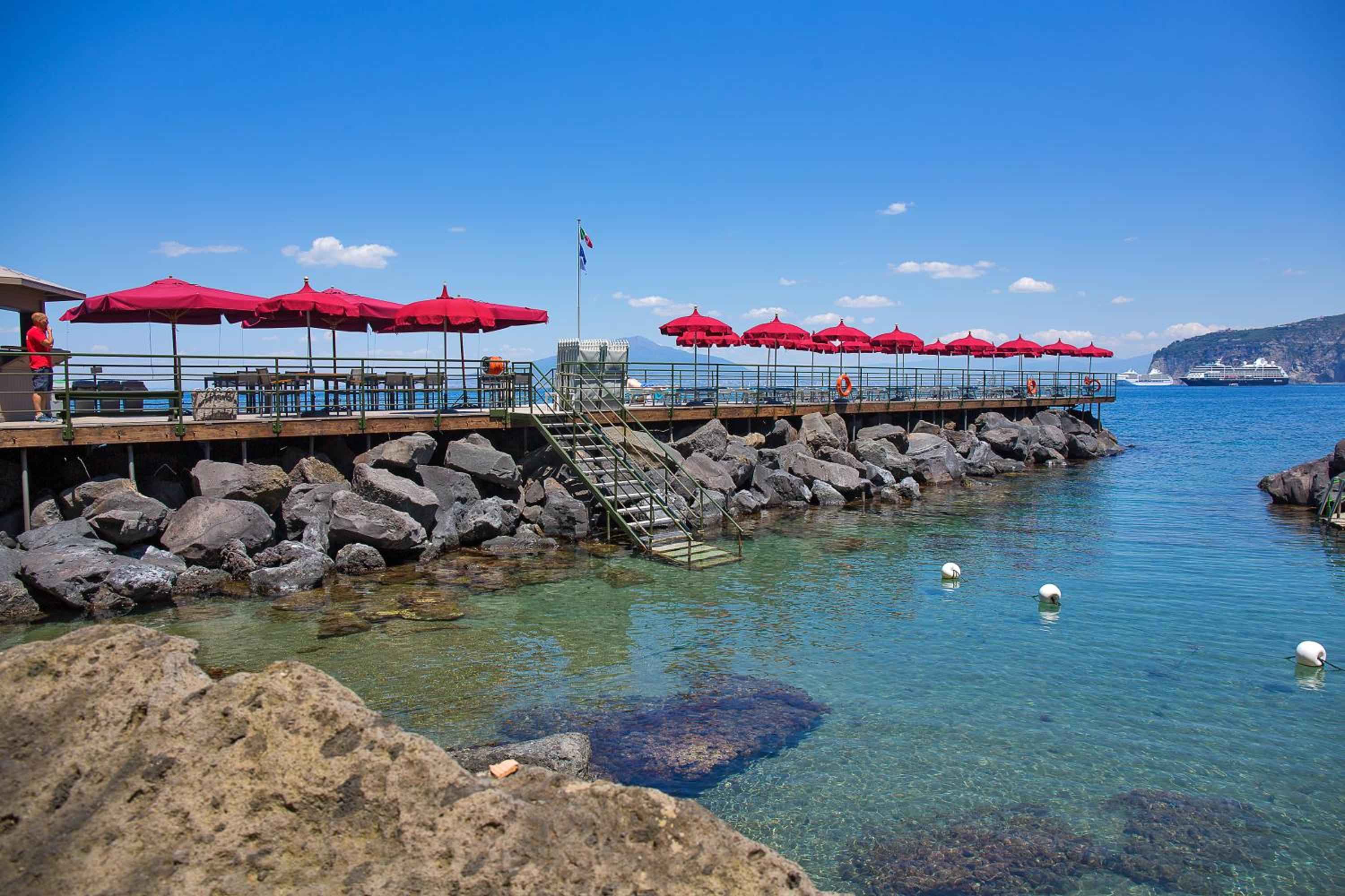 Red umbrellas dot the private pier where crystal-clear waters reveal rocky seabeds below, while cruise ships drift lazily across the azure Mediterranean horizon.