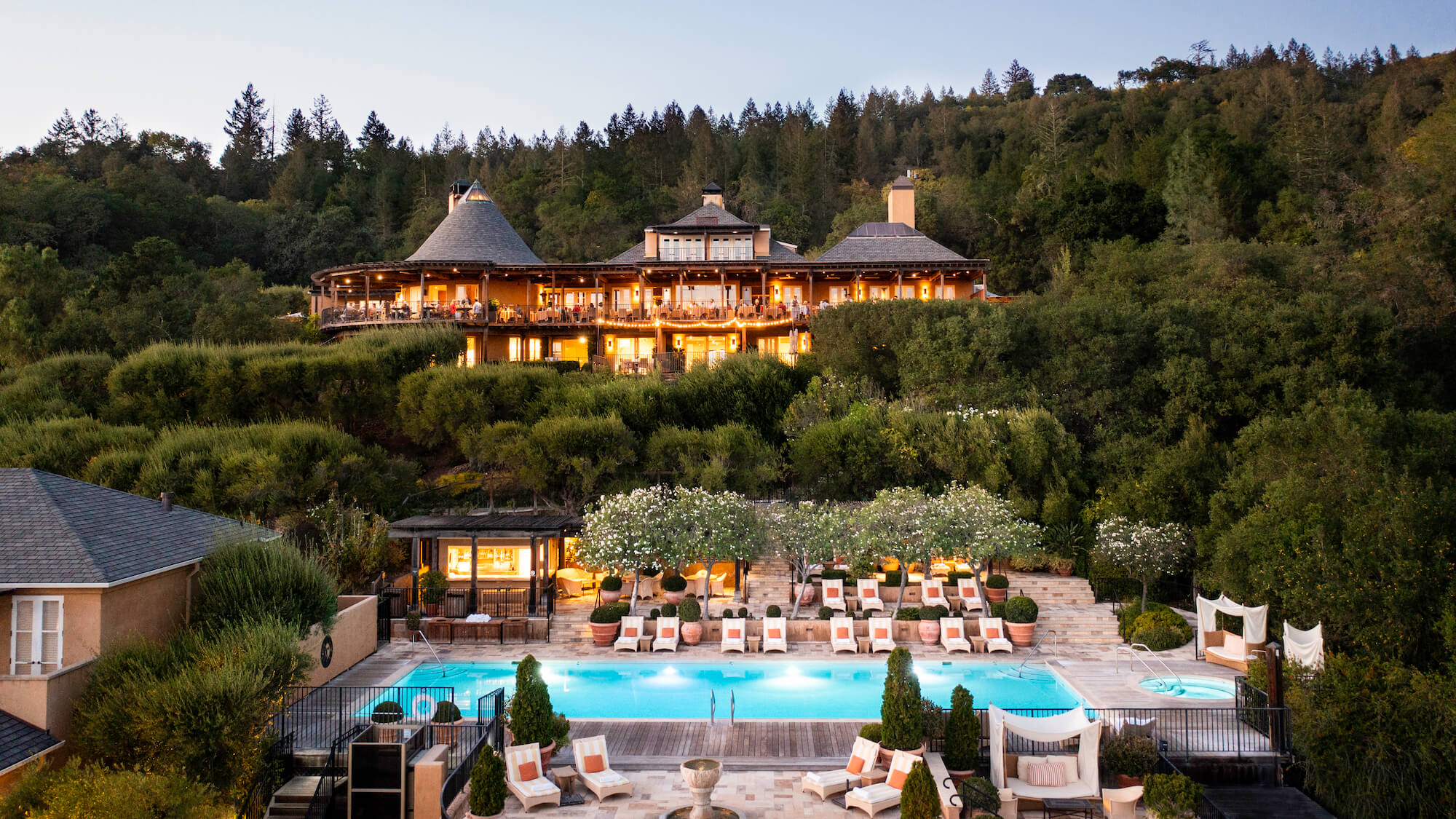 Terraced poolside loungers overlook vine-covered hillsides while the main lodge glows warmly against Napa Valley's forested ridgelines at golden hour.
