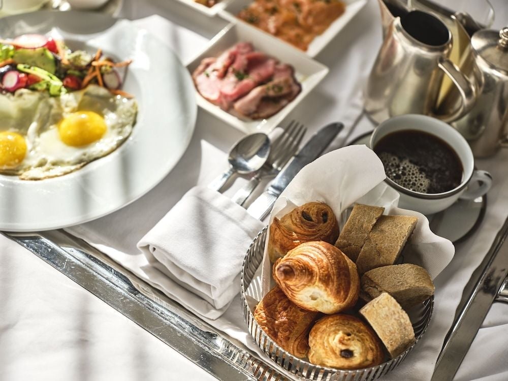Morning light illuminates a perfectly arranged breakfast spread—buttery pastries nestled in wicker, aromatic coffee steaming, and farm-fresh eggs glistening alongside vibrant greens.
