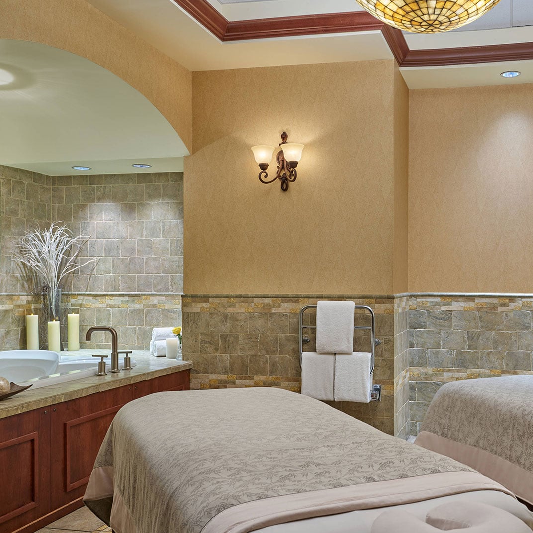 Warm stone walls and soft lighting create an intimate sanctuary where dual massage tables await beneath coffered ceilings adorned with ornate bronze sconces.