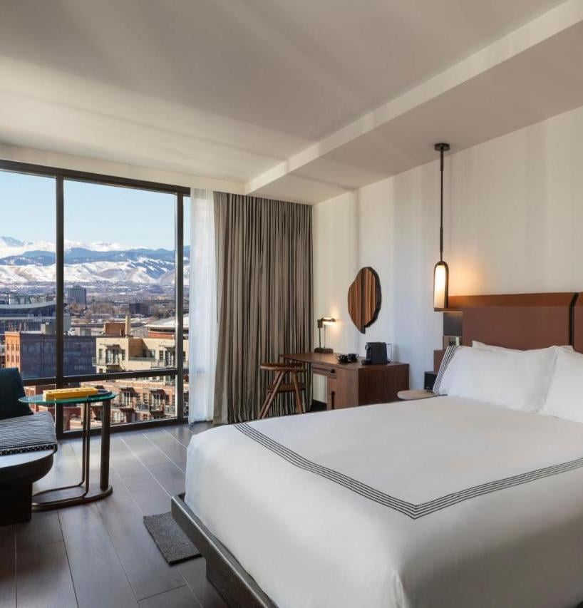Floor-to-ceiling windows frame snow-capped mountain vistas beyond downtown Denver, while warm wood tones and crisp white linens create a sophisticated urban sanctuary.