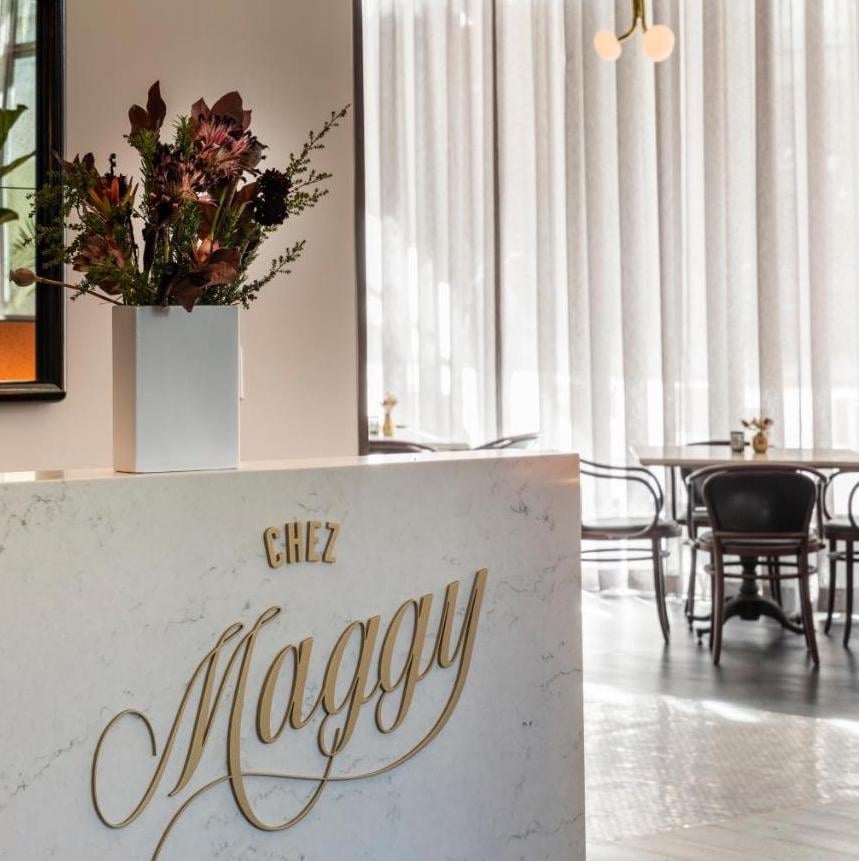 Chez Maggy's marble reception counter gleams beneath autumn florals, where golden script lettering hints at the French-inspired culinary romance awaiting beyond flowing white curtains.