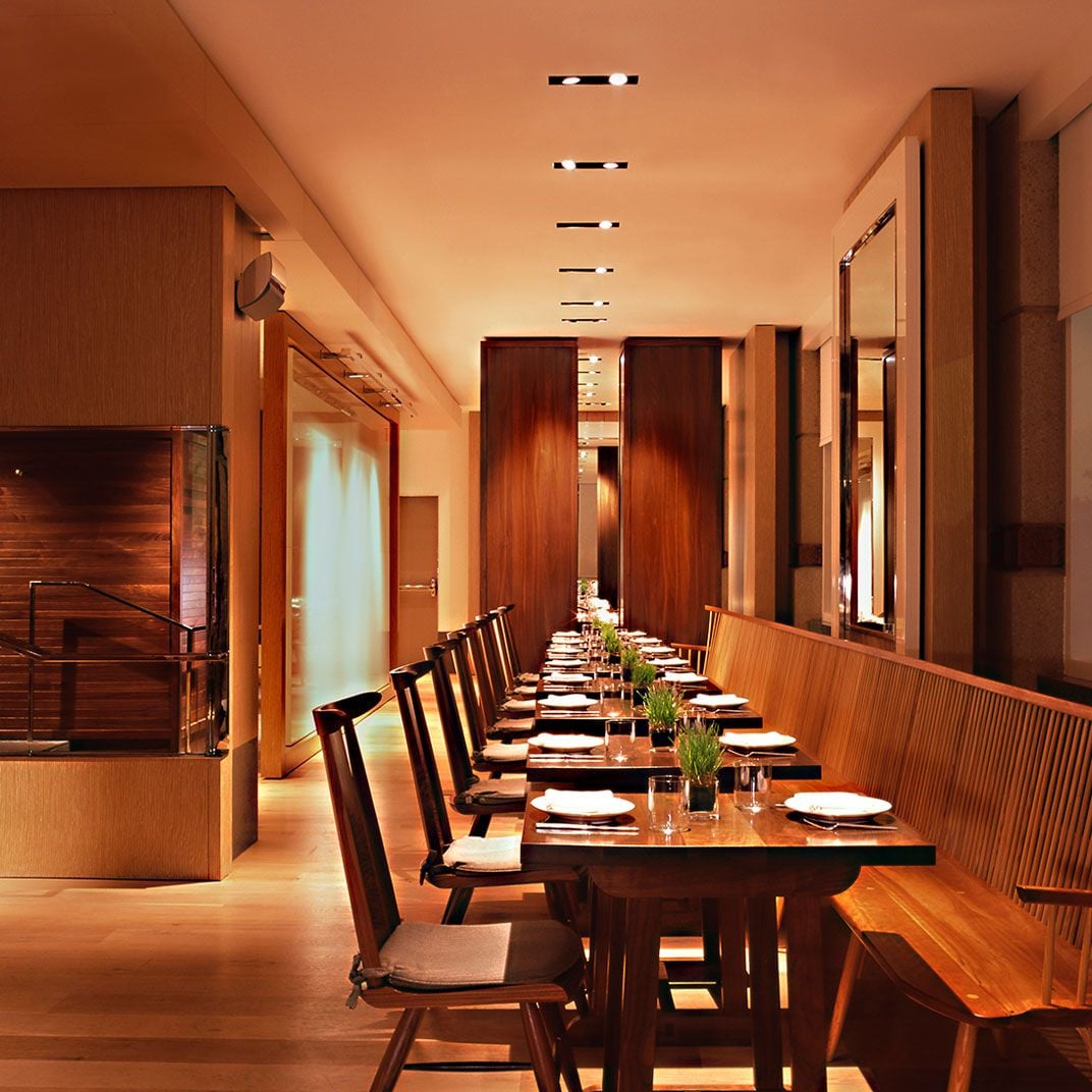 Warm wood paneling and amber lighting create an intimate dining atmosphere, where communal tables foster conversation beneath sleek recessed fixtures and rich mahogany accents.