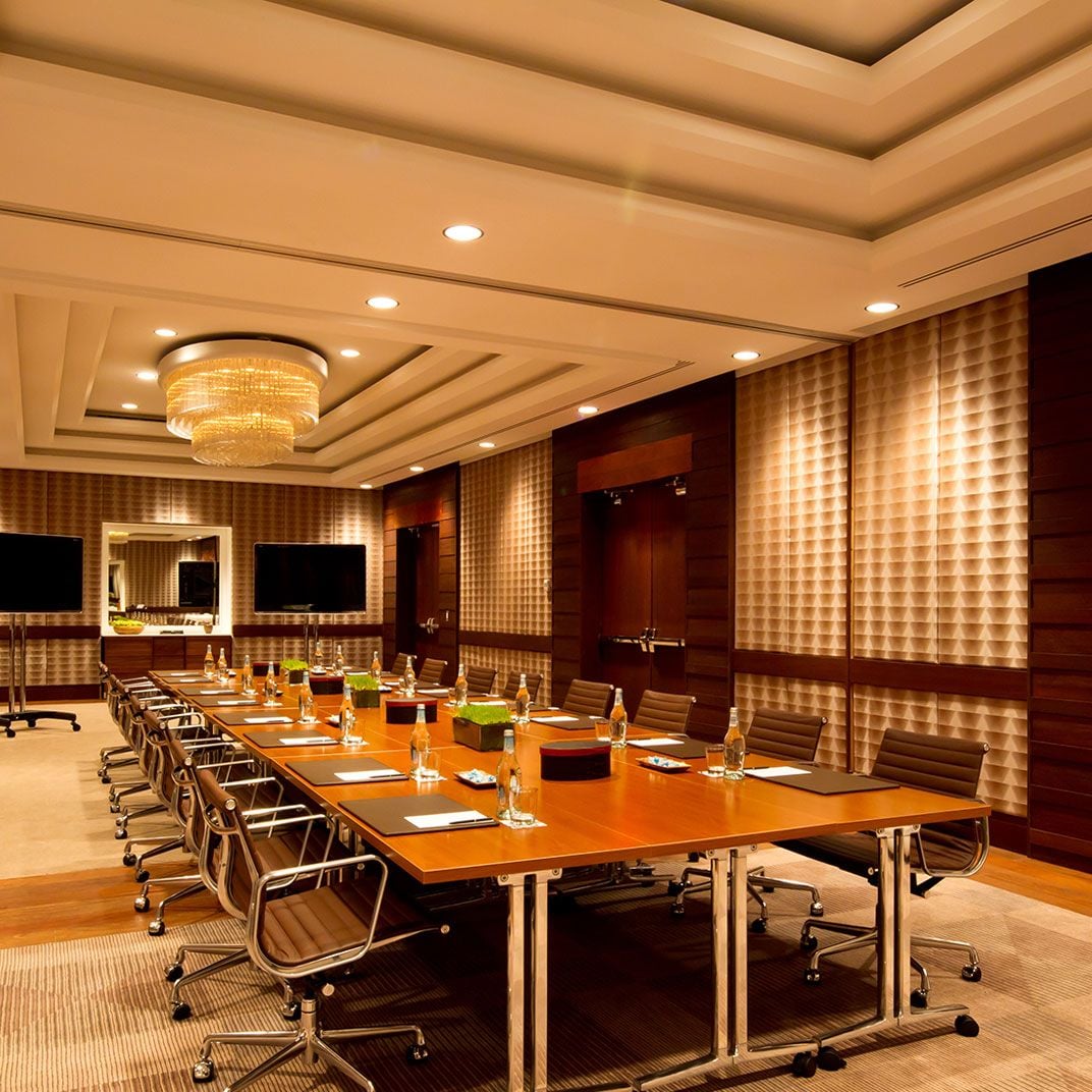 Golden chandeliers cast warm light across the boardroom's polished table, while textured wall panels and recessed lighting create an atmosphere of refined corporate elegance.