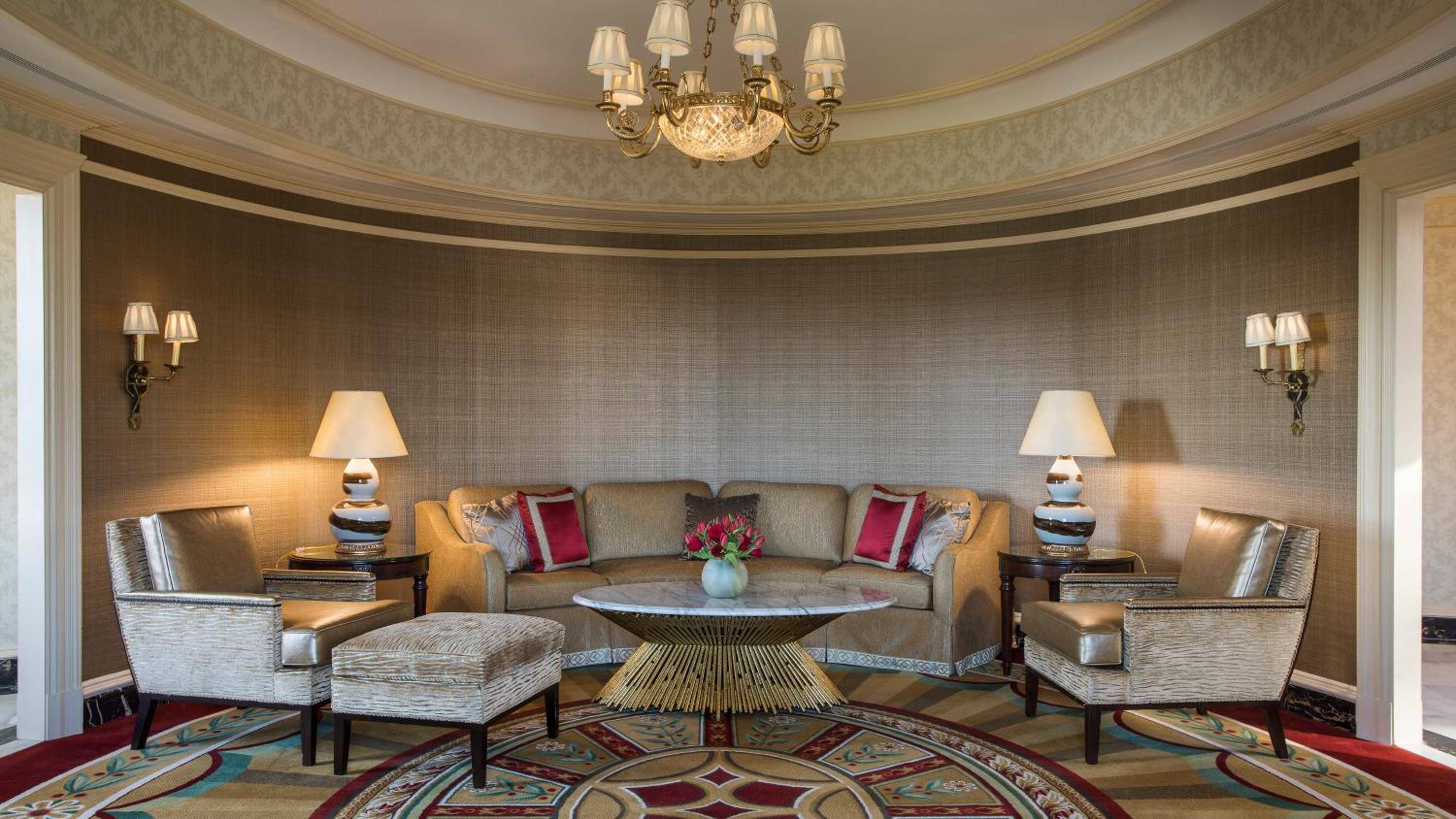 Warm lamplight illuminates this elegant curved sitting area, where neutral fabrics and rich textures create an intimate retreat beneath ornate moldings and a crystal chandelier.
