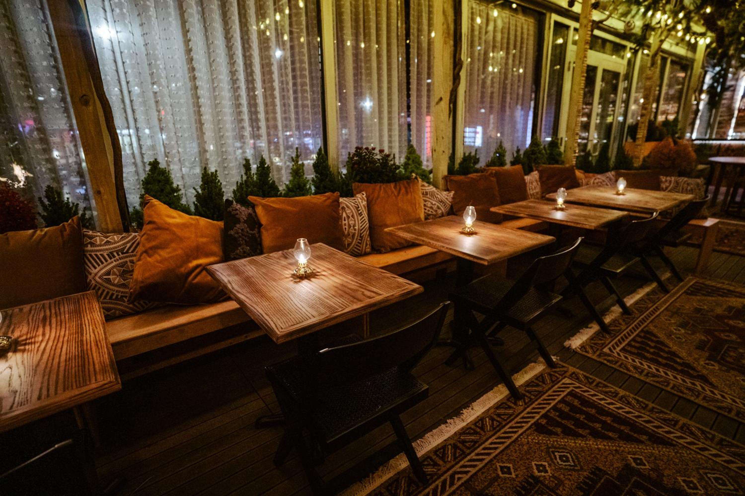 Warm amber lighting and geometric-patterned cushions create an intimate dining atmosphere, where string lights dance against floor-to-ceiling windows overlooking the bustling city beyond.