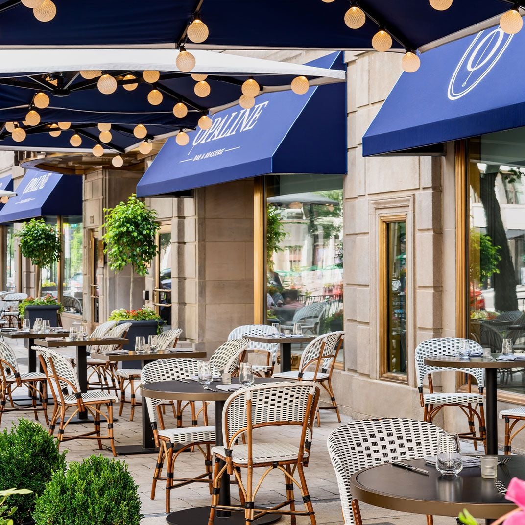 Parisian-style bistro chairs and glowing string lights create an enchanting al fresco dining scene beneath blue awnings, where French elegance meets Washington sophistication.