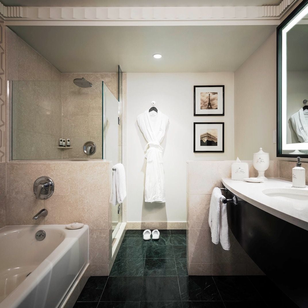 Marble surfaces gleam under soft lighting while plush robes await guests in this sophisticated bathroom, where Parisian elegance meets modern comfort through thoughtful amenities and refined finishes.