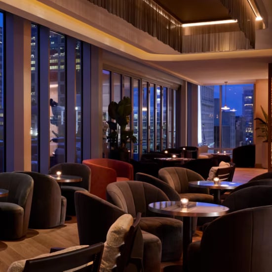 Plush velvet chairs and intimate lighting create a sophisticated evening retreat, while floor-to-ceiling windows frame Indianapolis's glittering skyline beyond warm wood paneling.