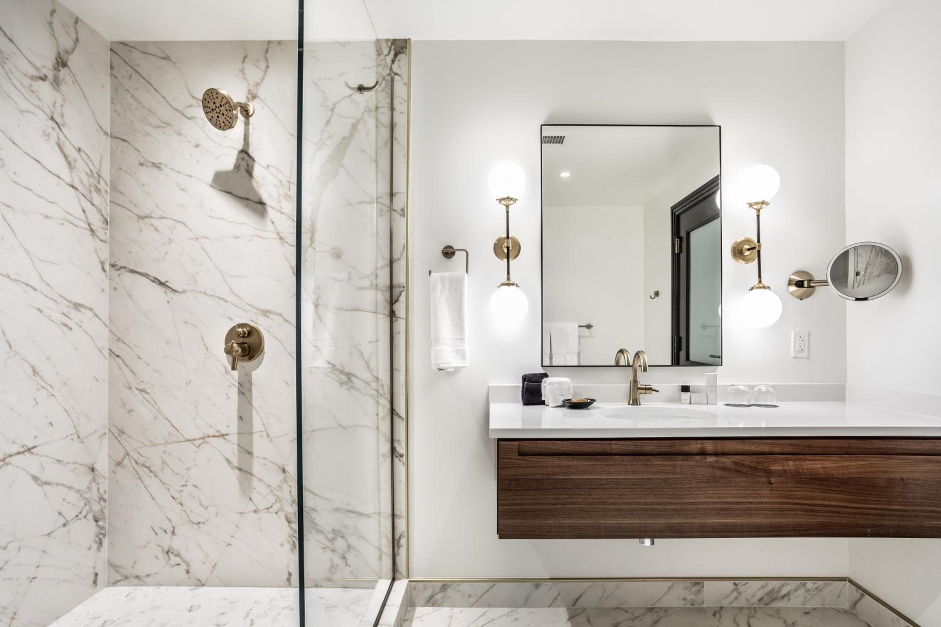 Carrara marble cascades across shower walls while warm brass fixtures and walnut vanity create an elevated sanctuary where modern luxury meets timeless sophistication.