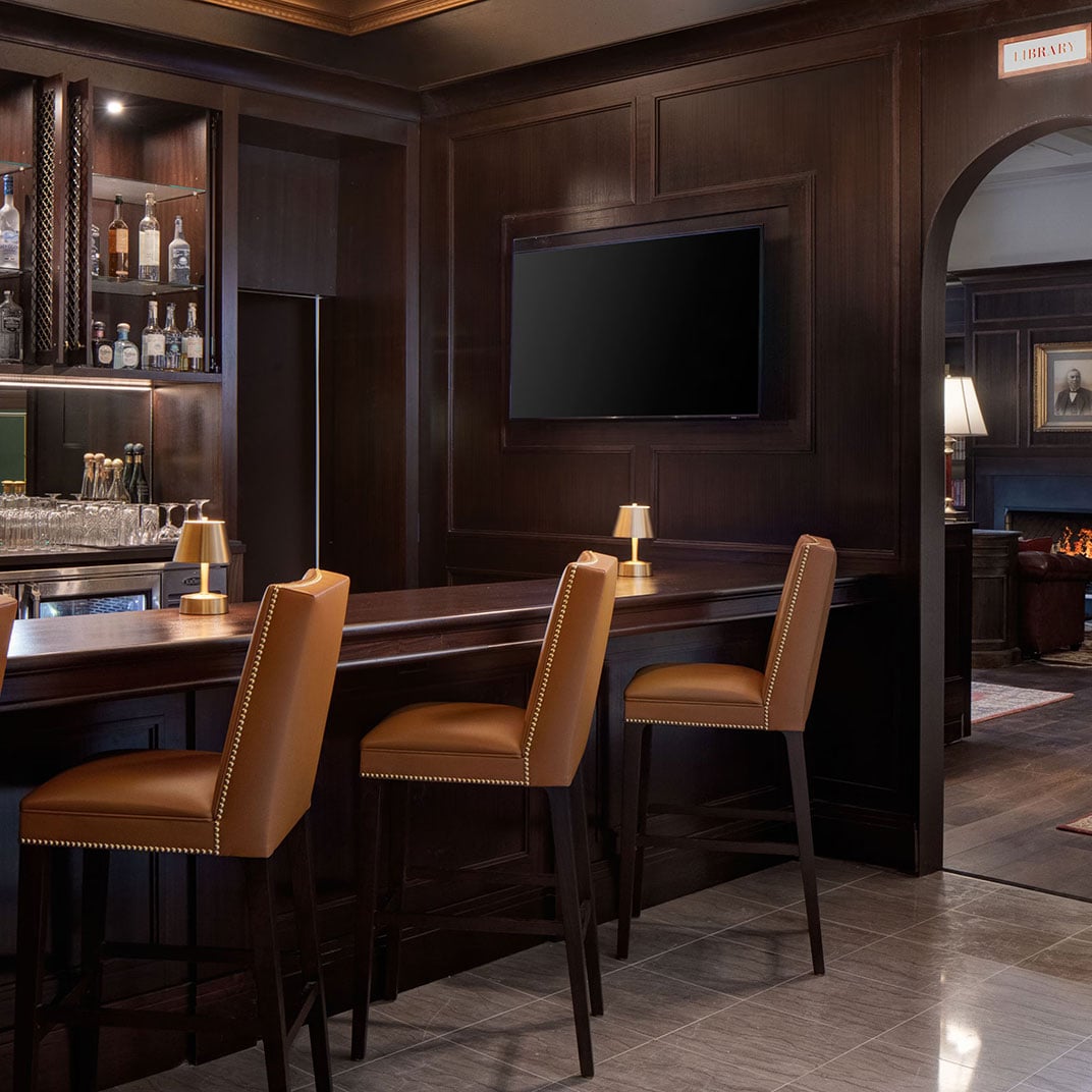 Rich walnut paneling and cognac leather stools create an intimate bar where crystal decanters catch warm light beneath the mounted screen.