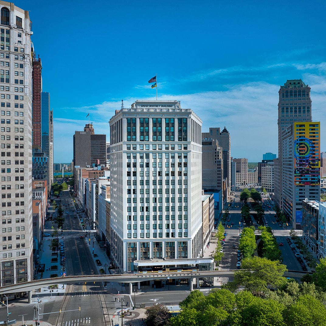 Downtown Detroit's grand David Whitney Building rises majestically above tree-lined streets, its historic limestone facade crowned with flags against brilliant blue Michigan skies.