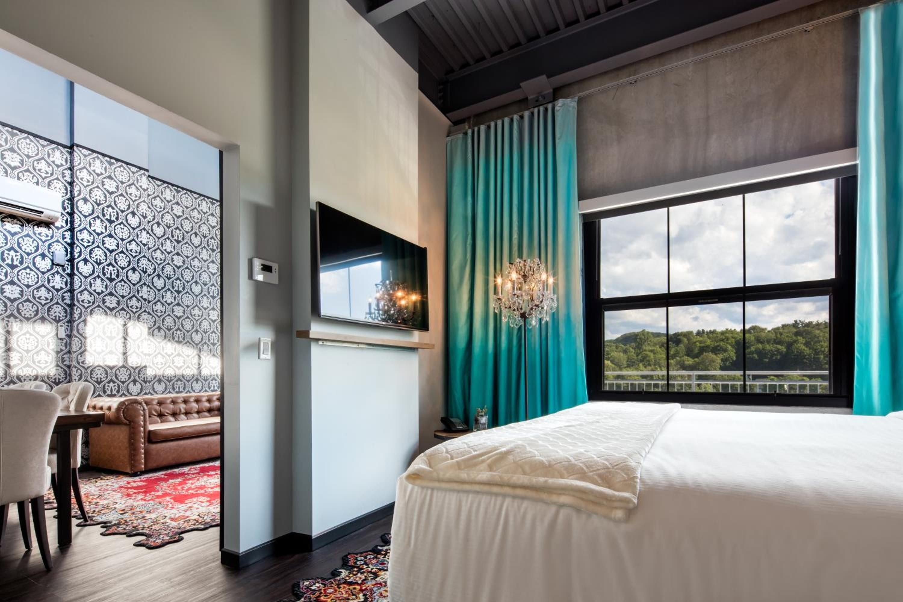 Turquoise silk curtains frame river views while exposed beams and patterned wallpaper create an eclectic sanctuary blending industrial chic with vintage elegance.