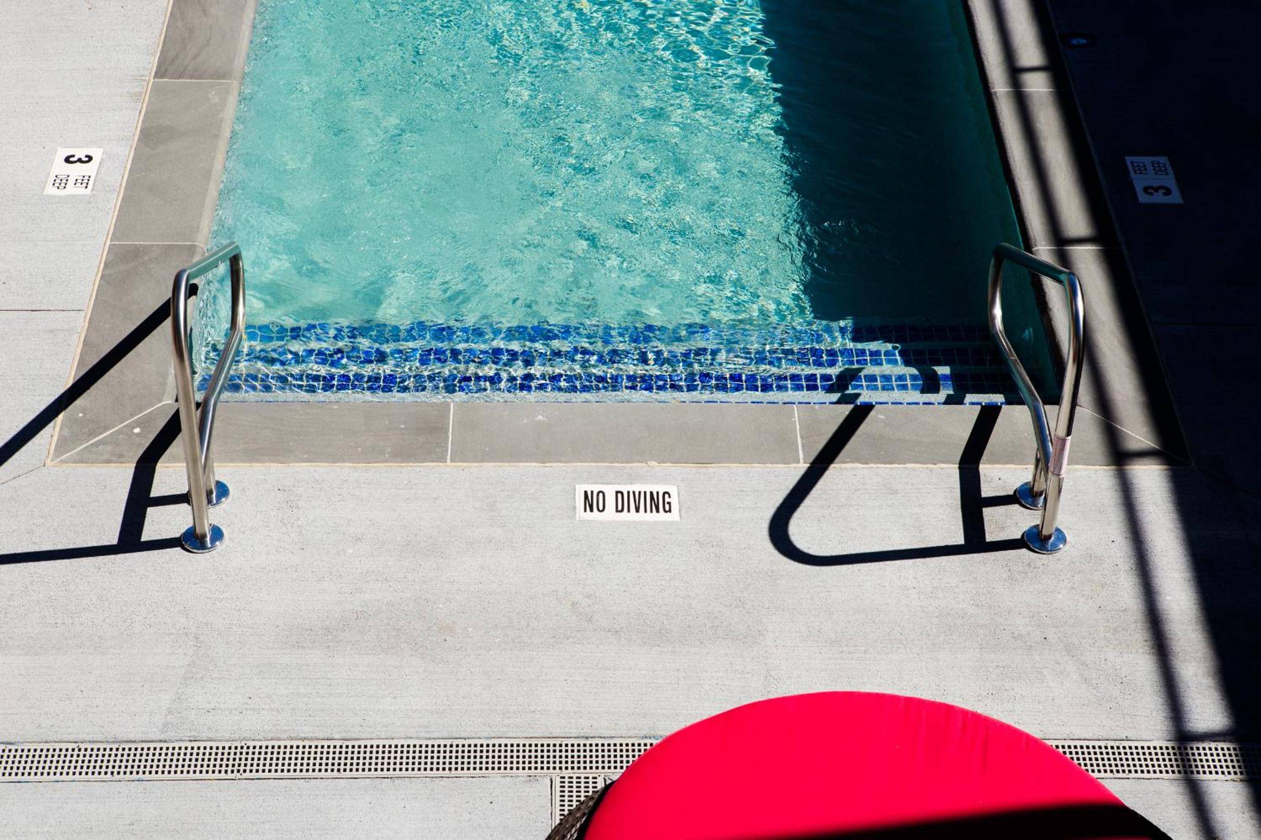 Turquoise waters meet mosaic tiles at the poolside entry, where chrome handrails frame a clear 'No Diving' reminder and shadows dance across sun-warmed concrete.