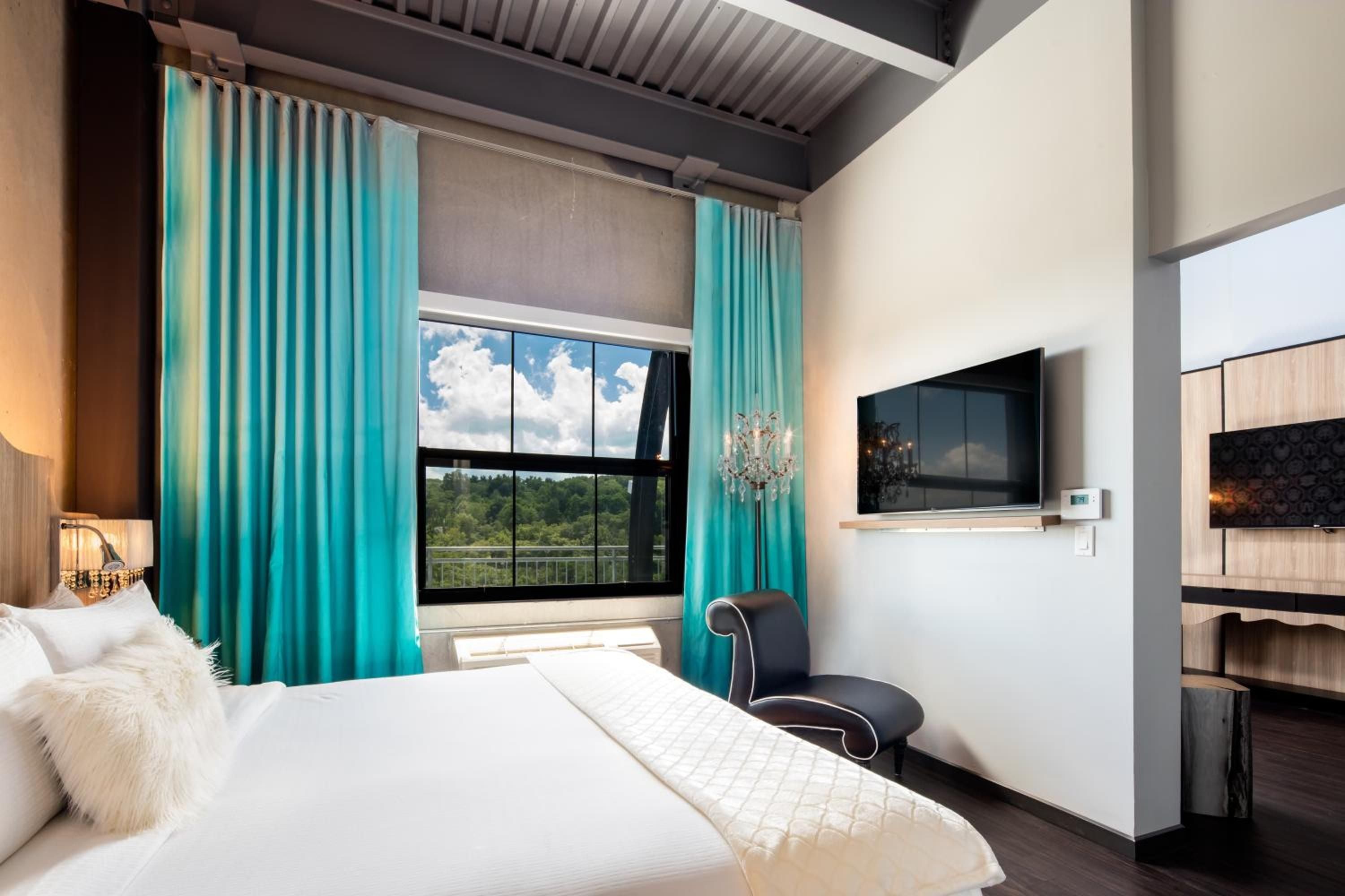 Vibrant turquoise curtains frame Hudson Valley views from this contemporary suite, where industrial ceiling details meet plush bedding and crystal chandelier elegance.