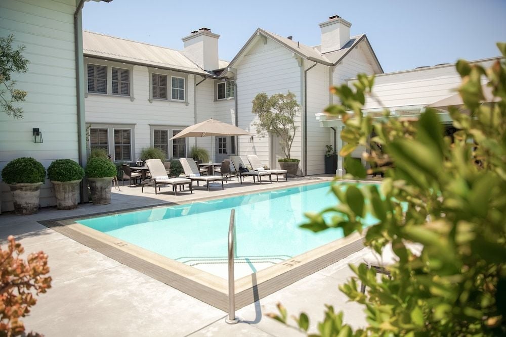 Lounge chairs frame the crystalline pool where California sun dances across water, while the whitewashed farmhouse architecture creates an intimate courtyard sanctuary.